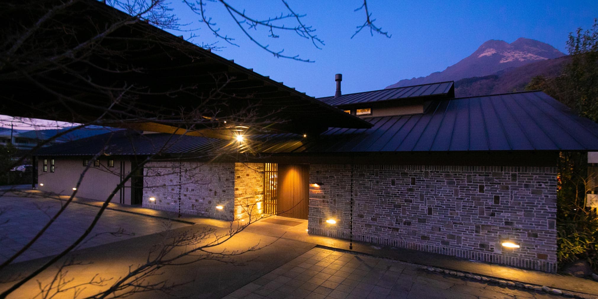 Yufuin Bettei Itsuki stone-and-tile exterior illuminated at dusk with Mount Yufu rising behind, Yufuin Onsen