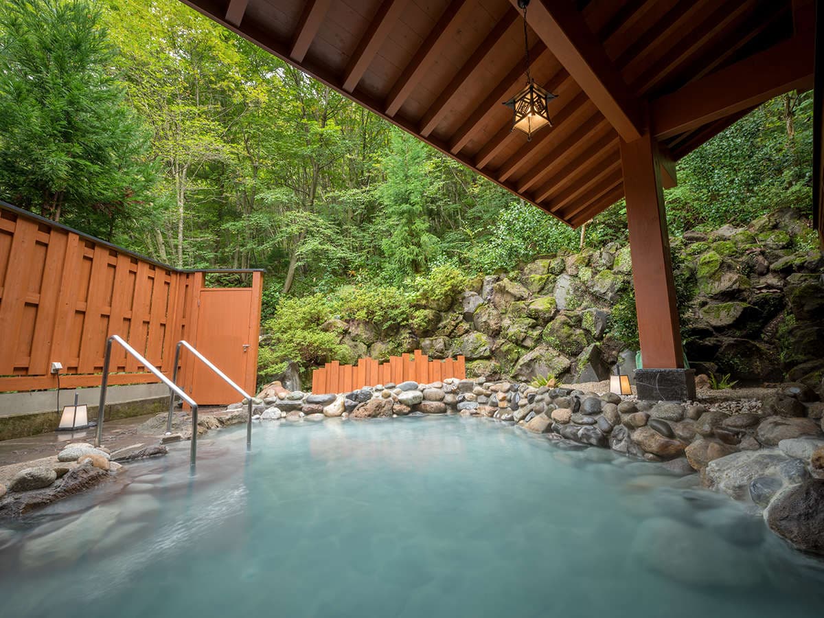 Open-air rotenburo bath surrounded by mossy boulders and timber fencing under a wooden eave, Yumoto Kissho, Naruko Onsen