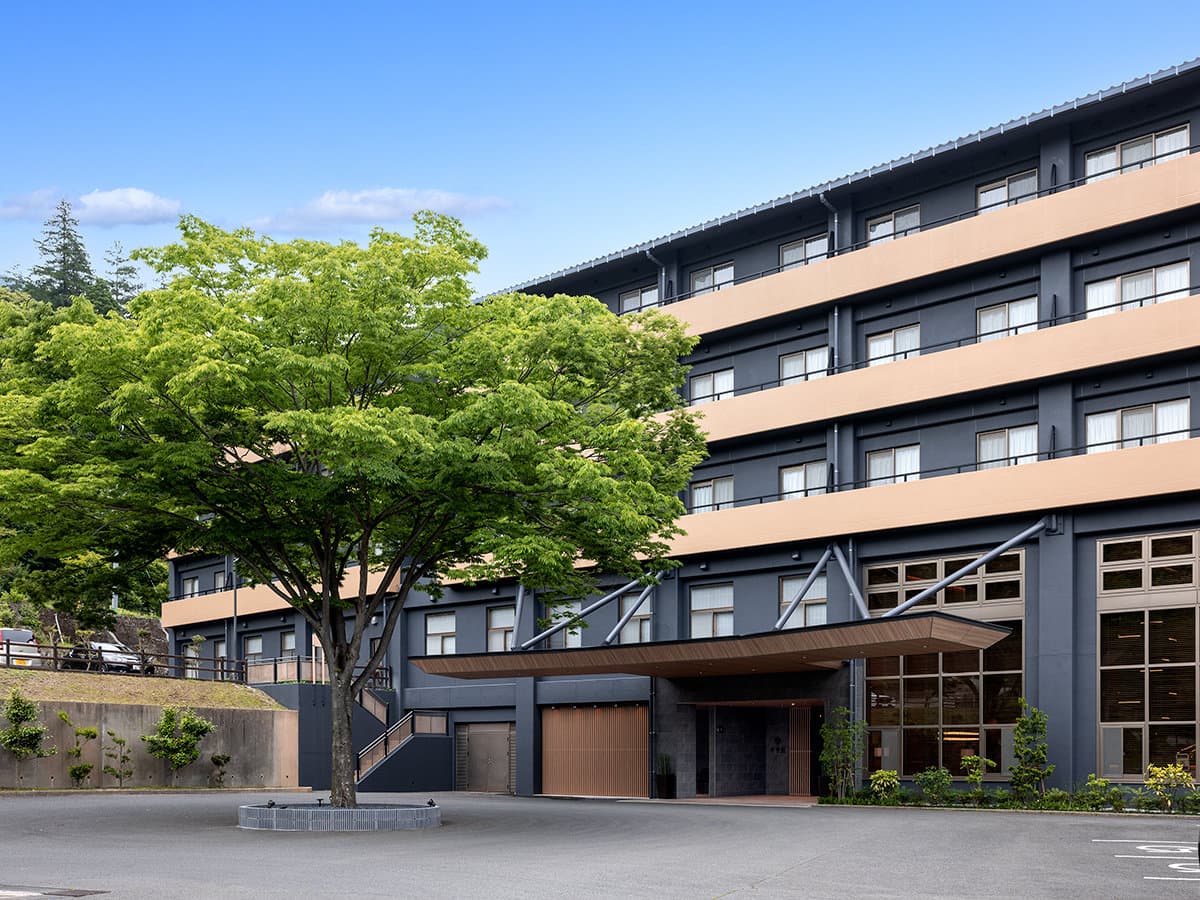 Daytime exterior of Seiunso in Unzen Onsen, dark facade with wooden balconies beside a green maple tree