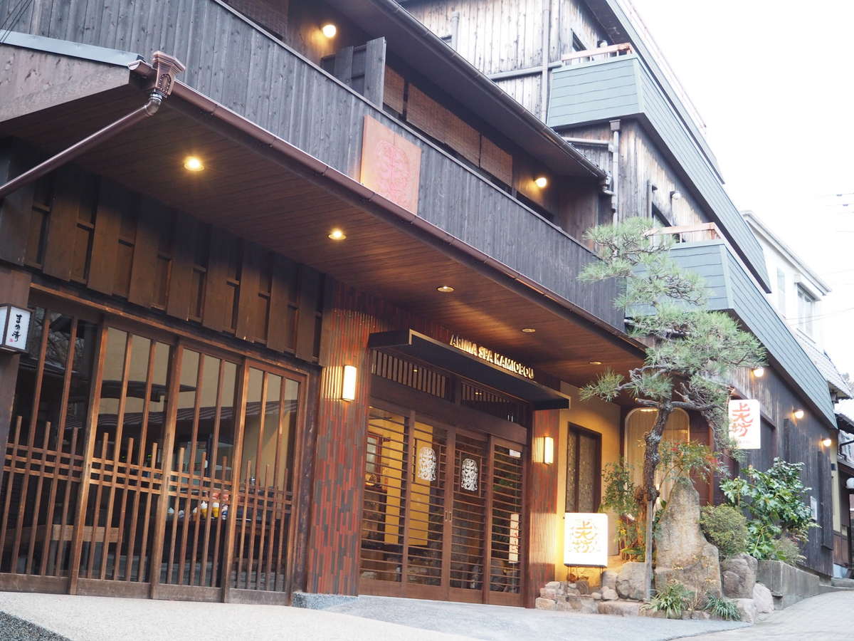 Kamiobo's traditional dark-timber ryokan facade with lantern-lit lattice entrance and a pine tree on a quiet Arima Onsen lane