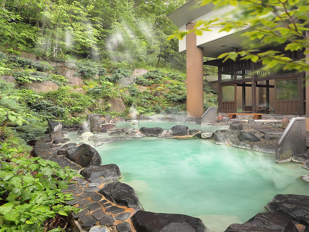 Milky white open-air rotenburo hot spring bath surrounded by rocks and green forest, steam rising at Zao Onsen