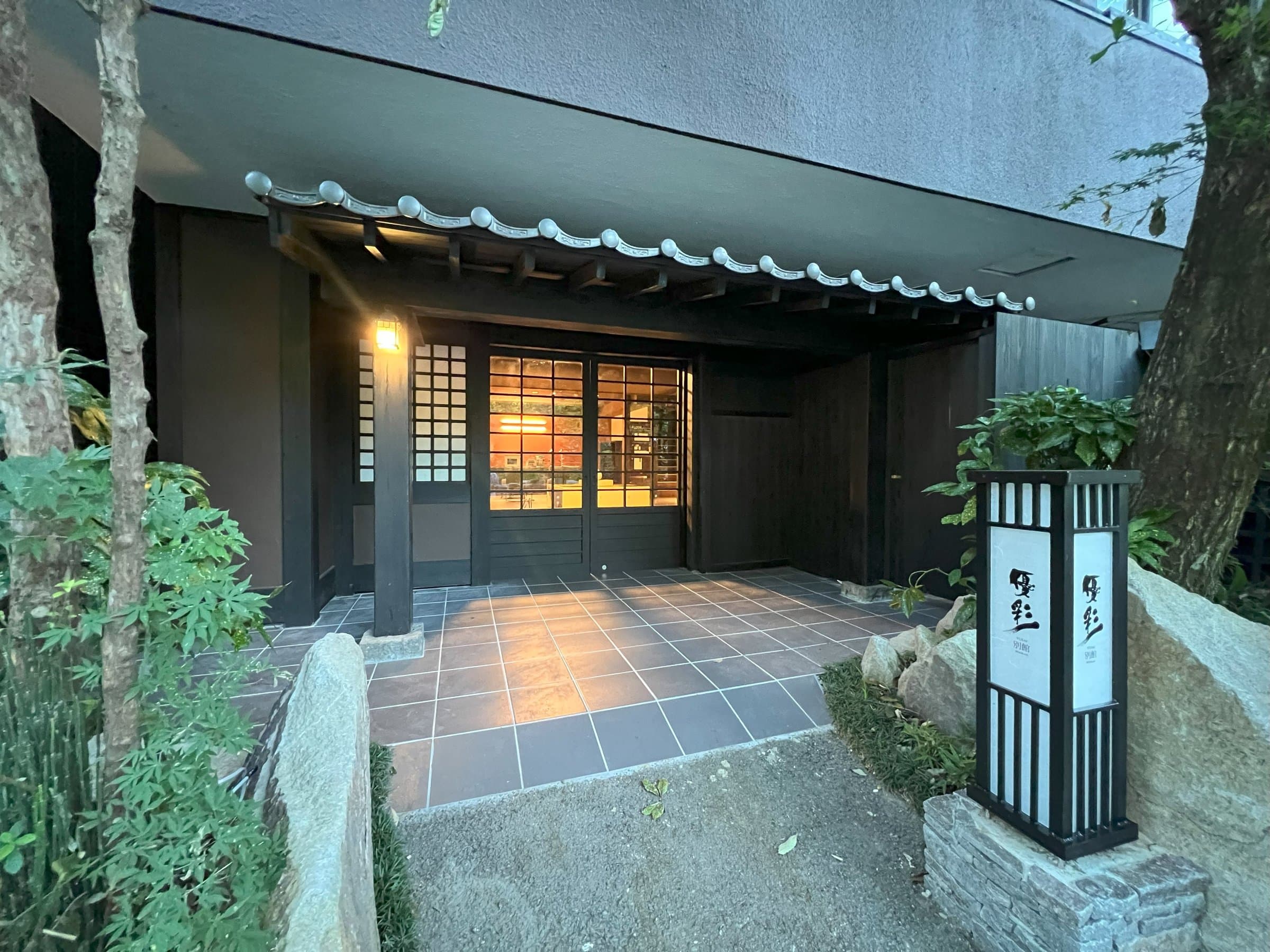 Yusai sukiya-style annex entrance gate with tiled roof, andon lantern and greenery at dusk