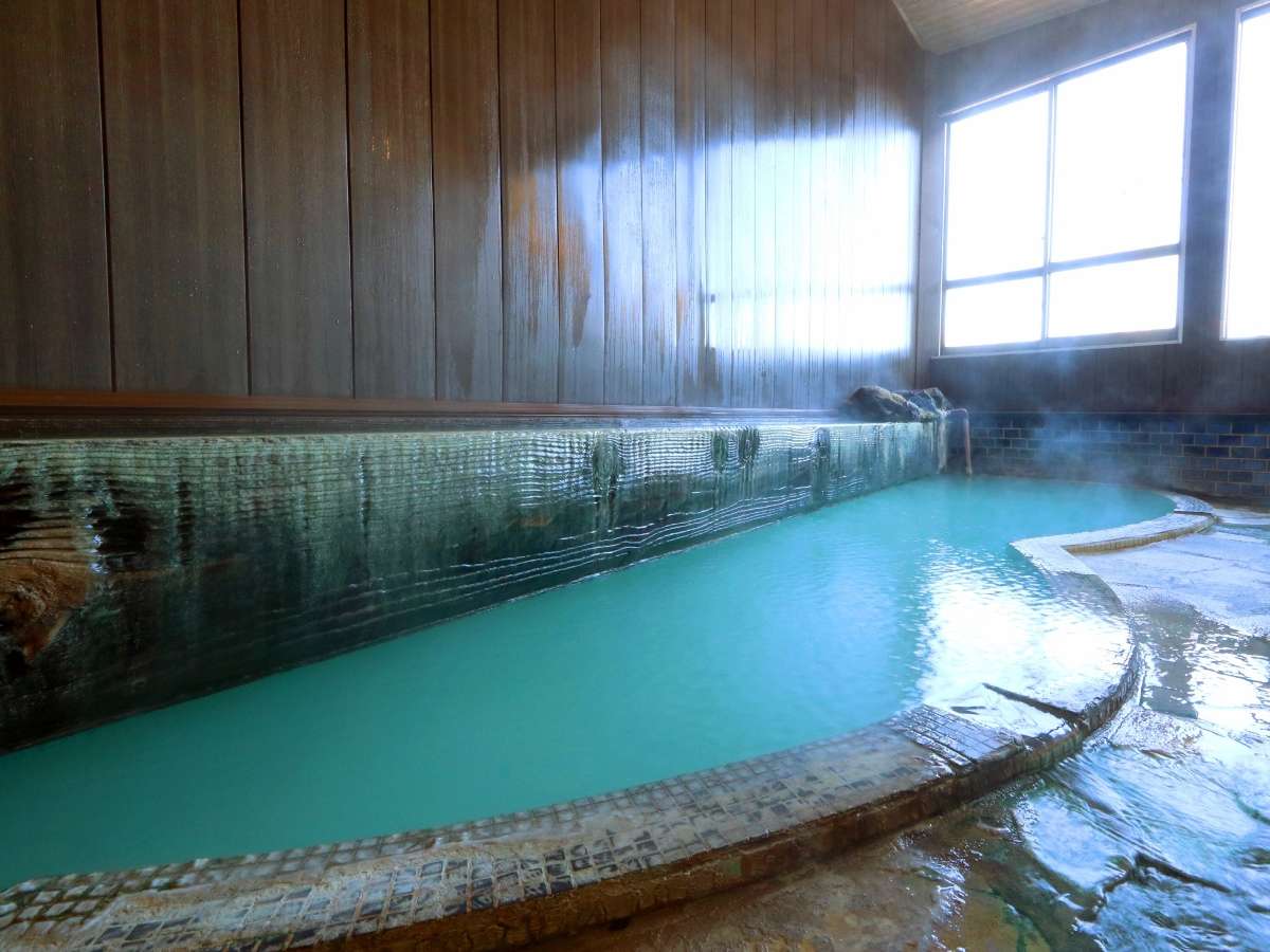 Indoor hot spring bath with milky turquoise water, wooden walls and rising steam by a bright window