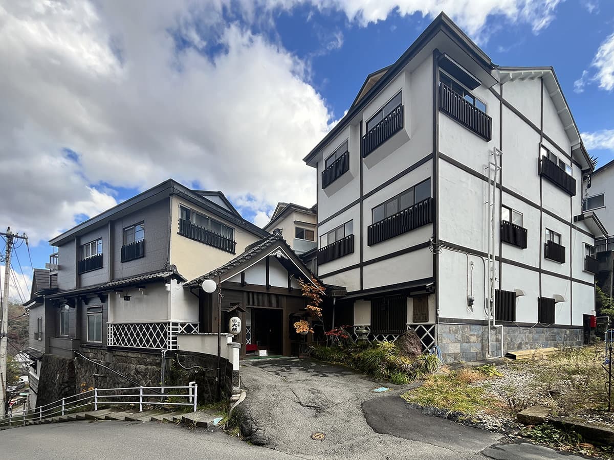 White-walled traditional Japanese inn exterior with tiered roofs and a tiled entrance on a hillside under blue sky
