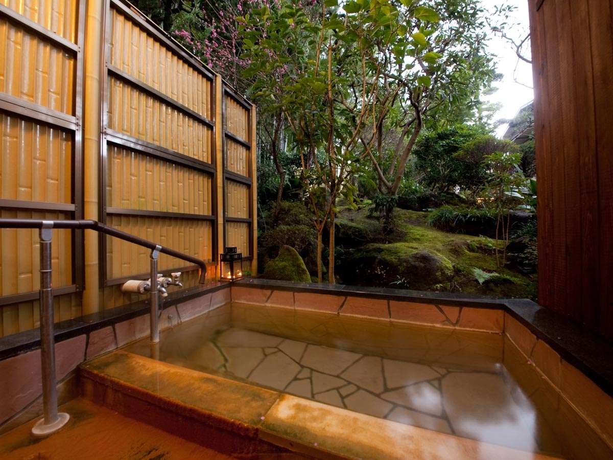Open-air stone bath behind a bamboo fence facing a mossy green garden with blossoms, Ryokan Sennari, Beppu Onsen