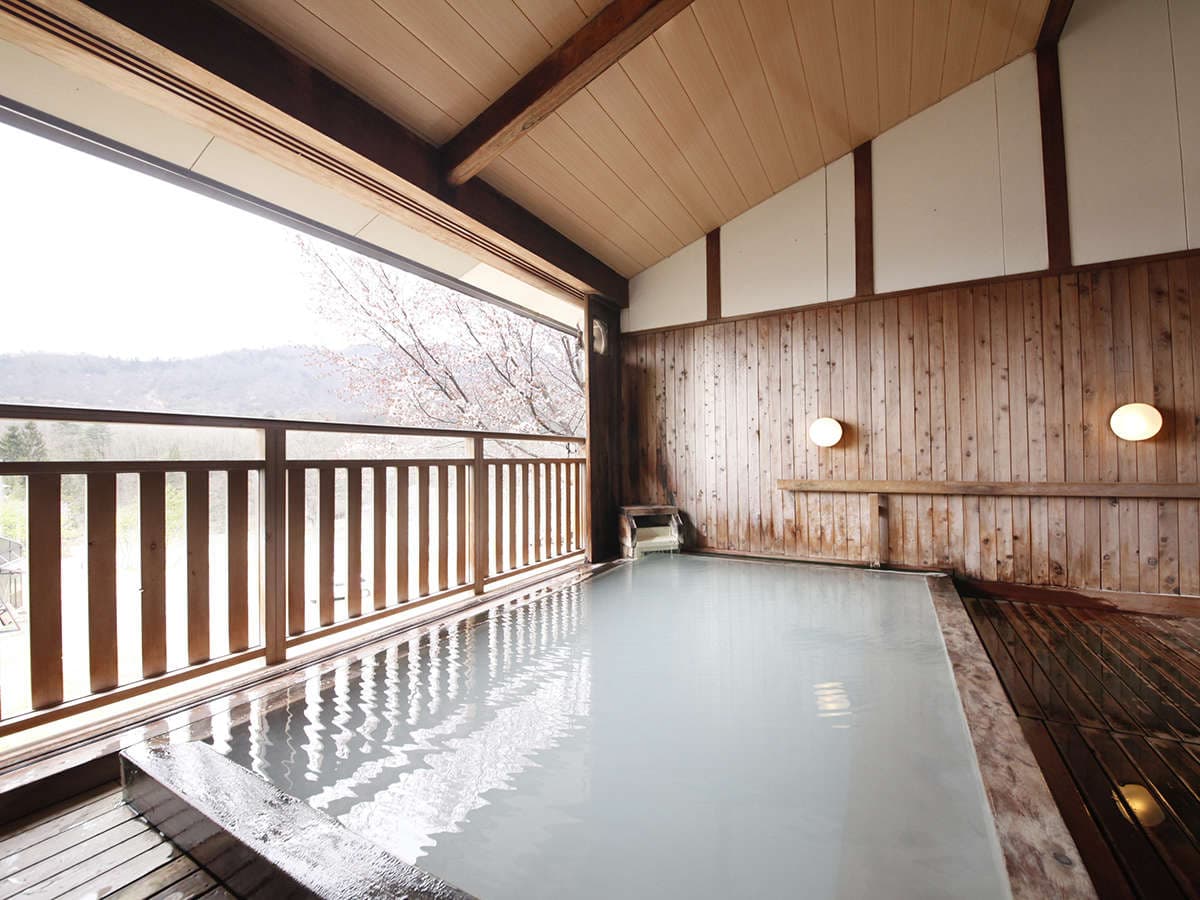 Open-air bath with cloudy hot-spring water framing blossoming trees and the Zao Onsen mountains