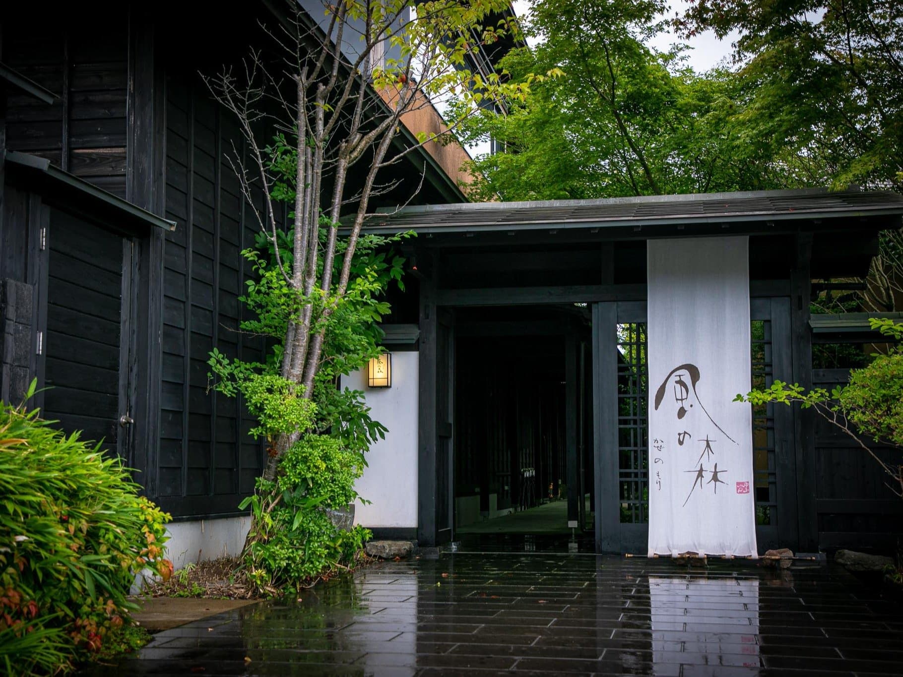 Dark timber facade and noren-curtained entrance of Yufuin Bettei Kazenomori ryokan after rain in Yufuin Onsen