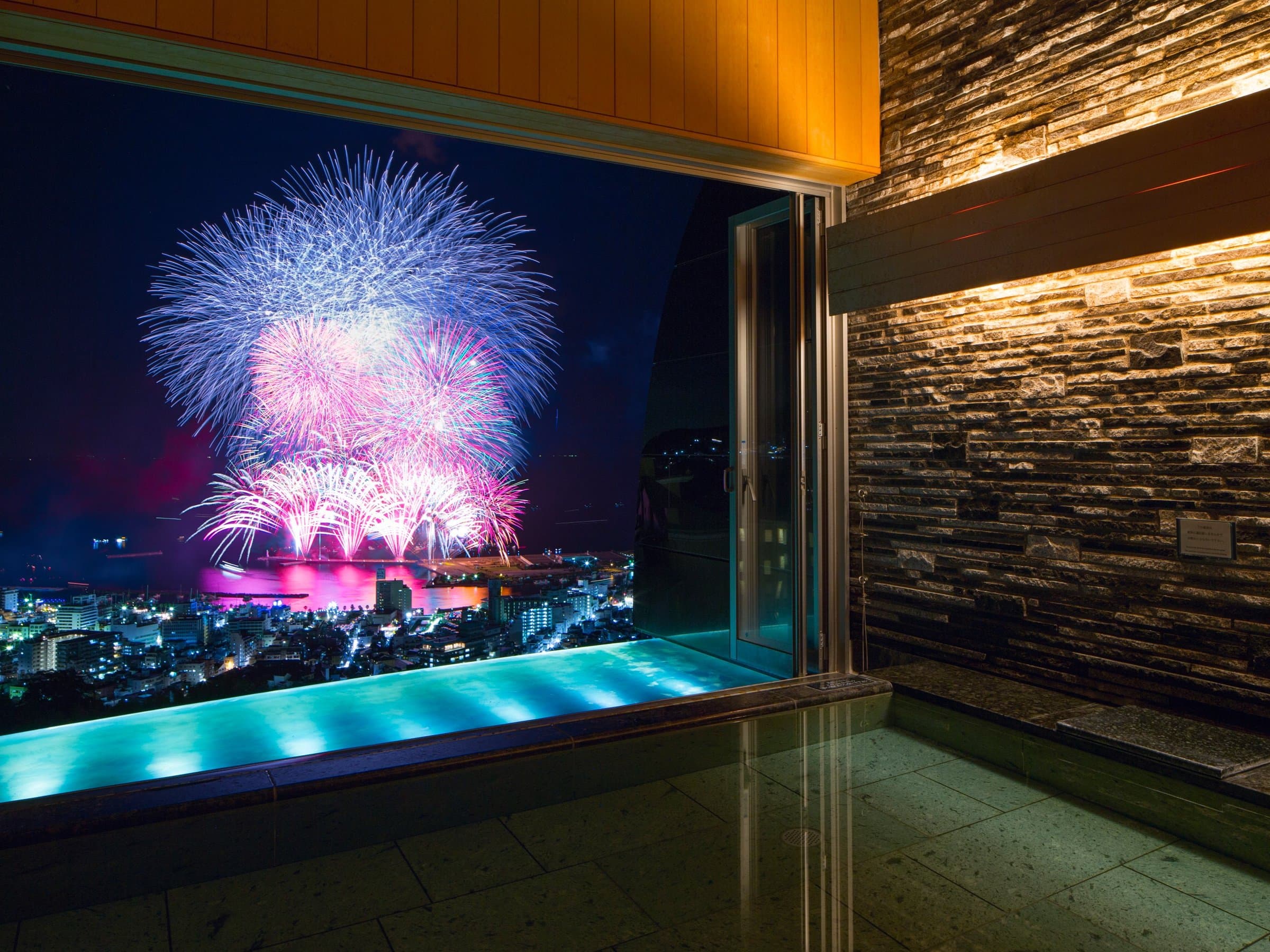 Open-air stone rotenburo bath at Risonare Atami framed by fireworks over Atami town at night