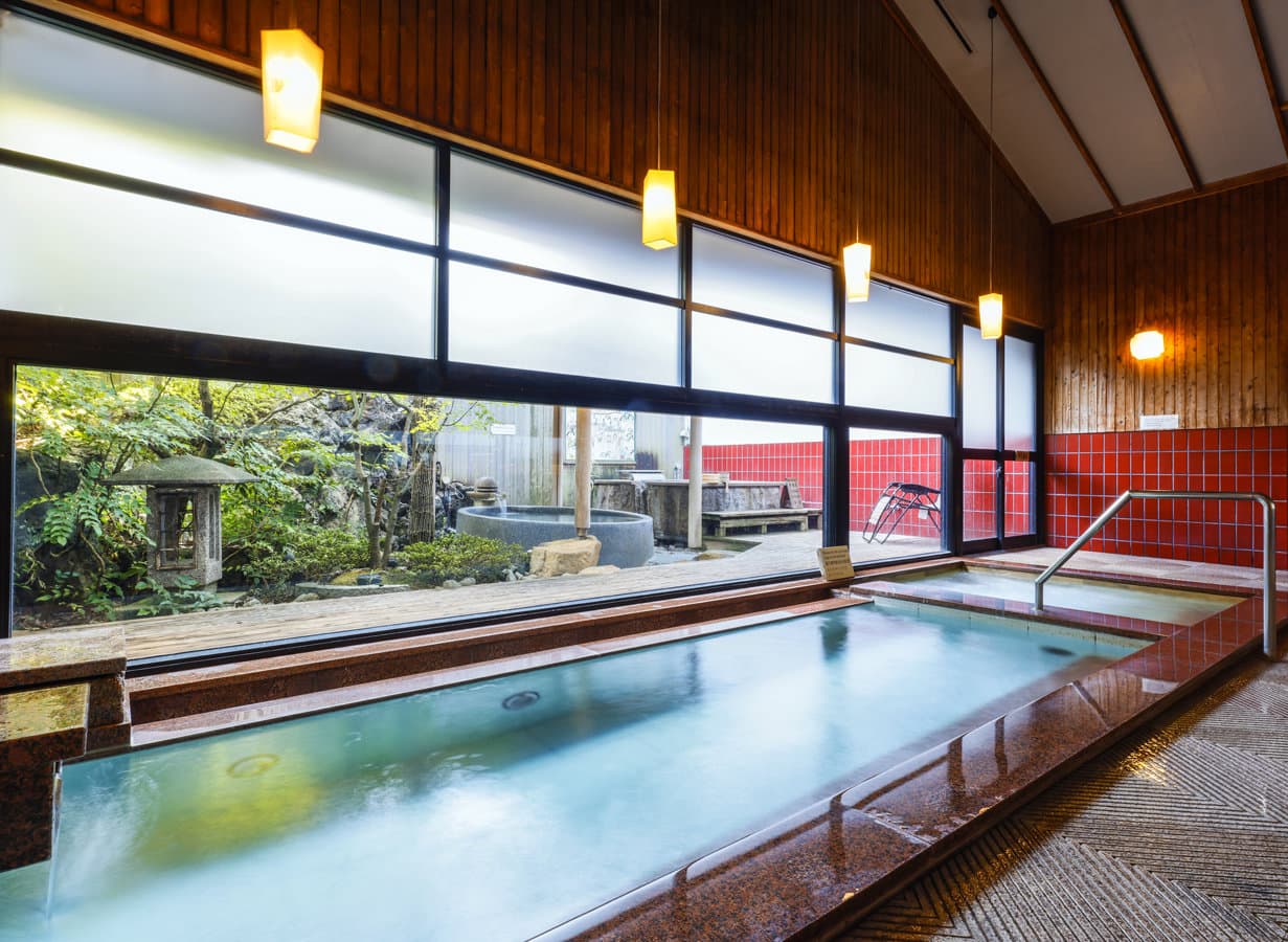 Indoor hot-spring bath in dark granite with hanging lanterns overlooking a rotenburo and garden, Onishiya Suishoen, Kinosaki Onsen