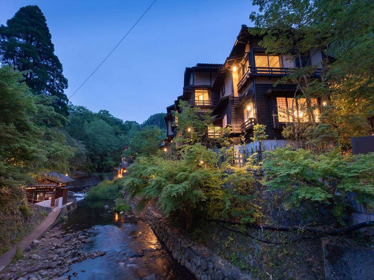 Kurokawa Onsen Fujiya ryokan's timber buildings rising above a rocky mountain stream at dusk with windows lit