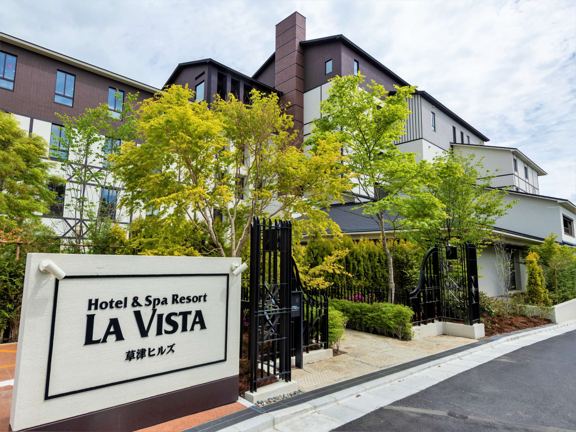 Entrance of Hotel & Spa Resort La Vista Kusatsu Hills with its signboard, black gate and dark-clad hillside building framed by greenery