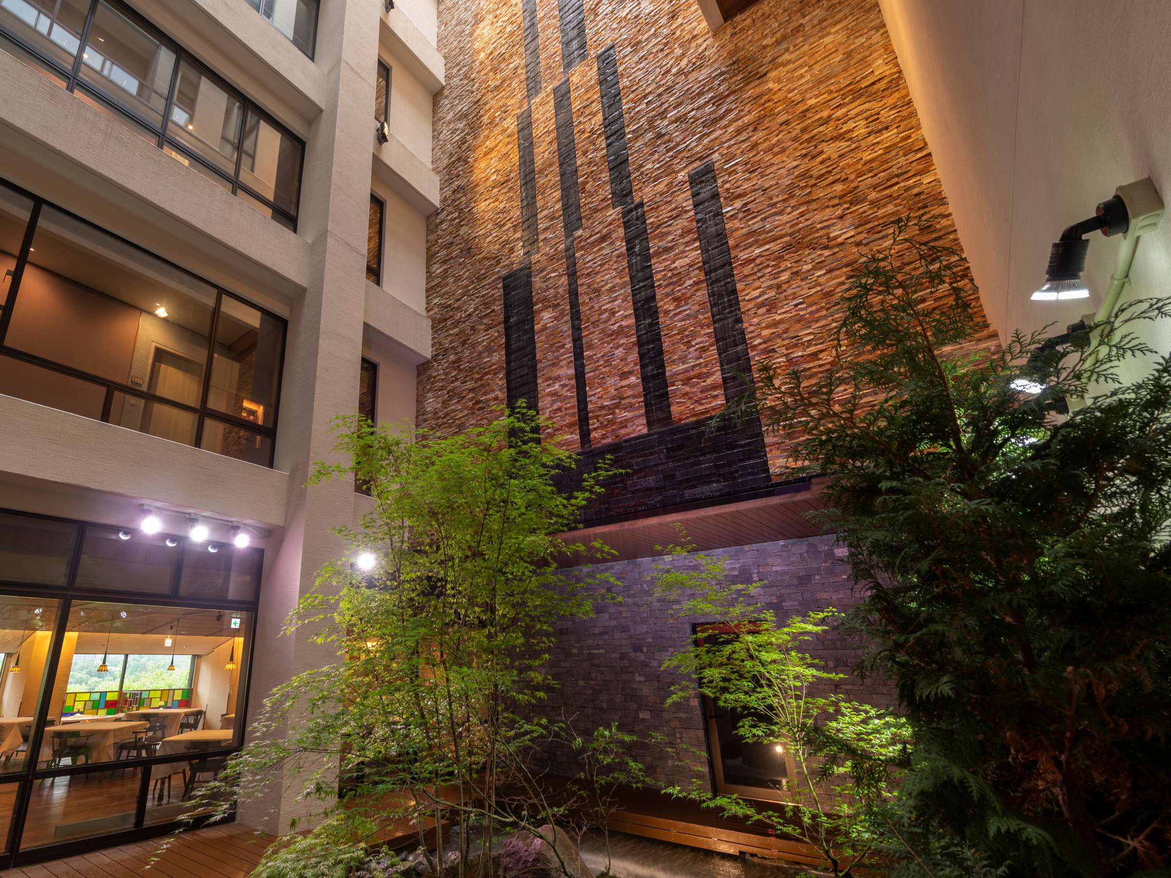 Illuminated stone-clad atrium courtyard of La Vista Kusatsu Hills at night with maple trees and a glowing dining room below