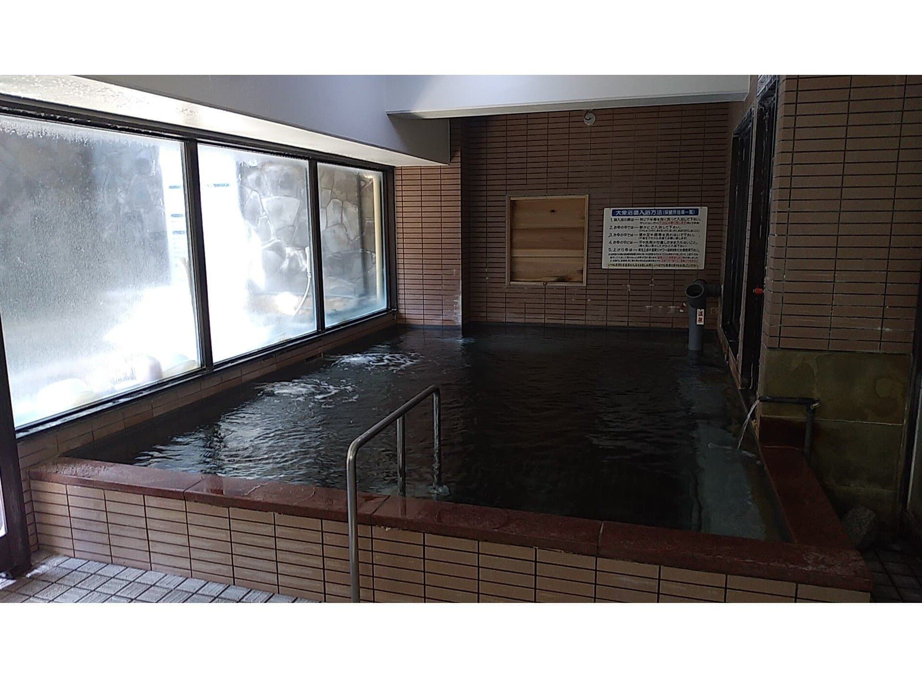 Indoor hot spring bath at Fuji-no-en with a tiled stone-edged pool and a view window onto the rock-lined outdoor bath