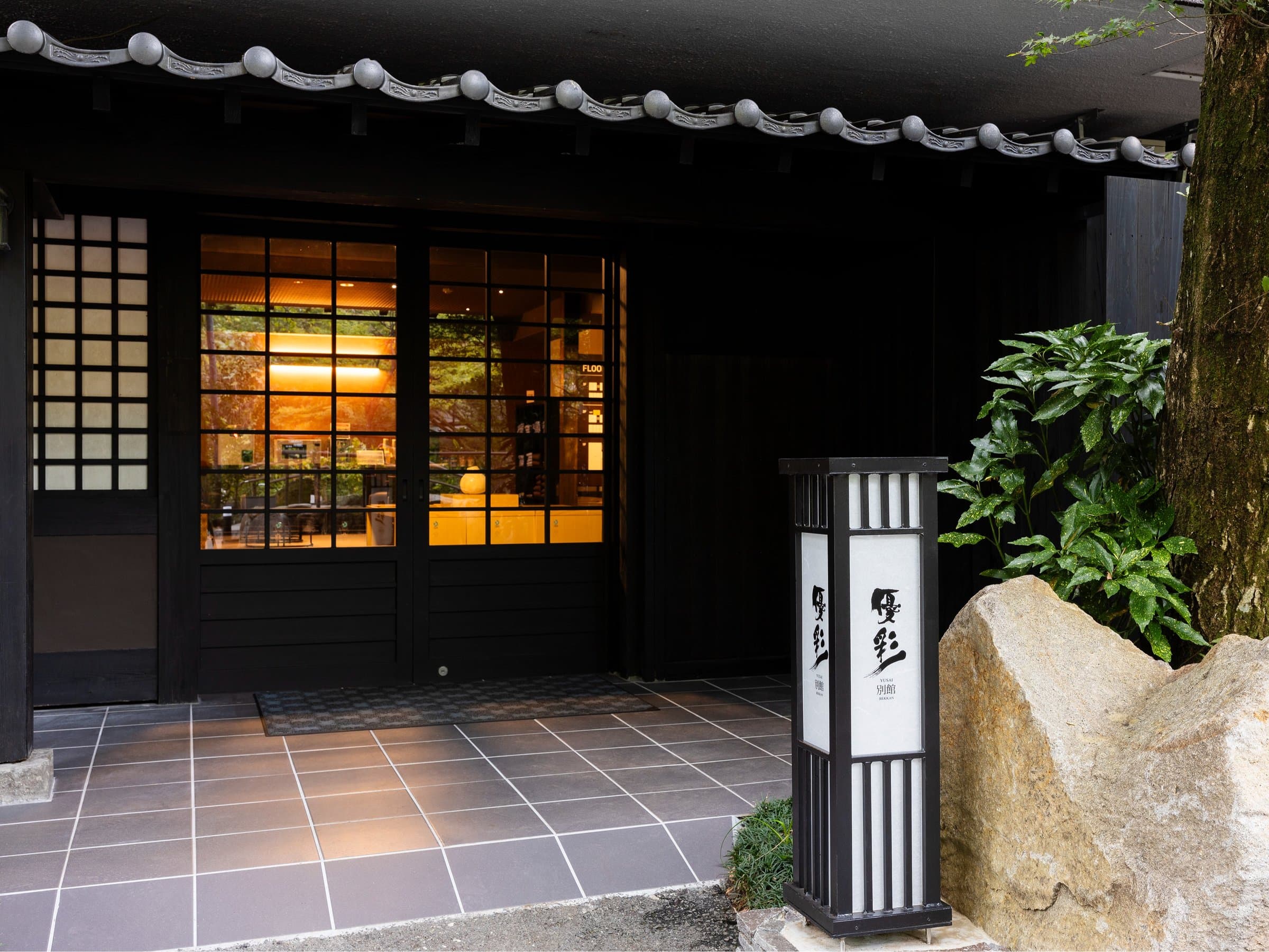 Yusai Bekkan front entrance at dusk, dark timber facade with lit interior and an illuminated 'Yusai' name lantern