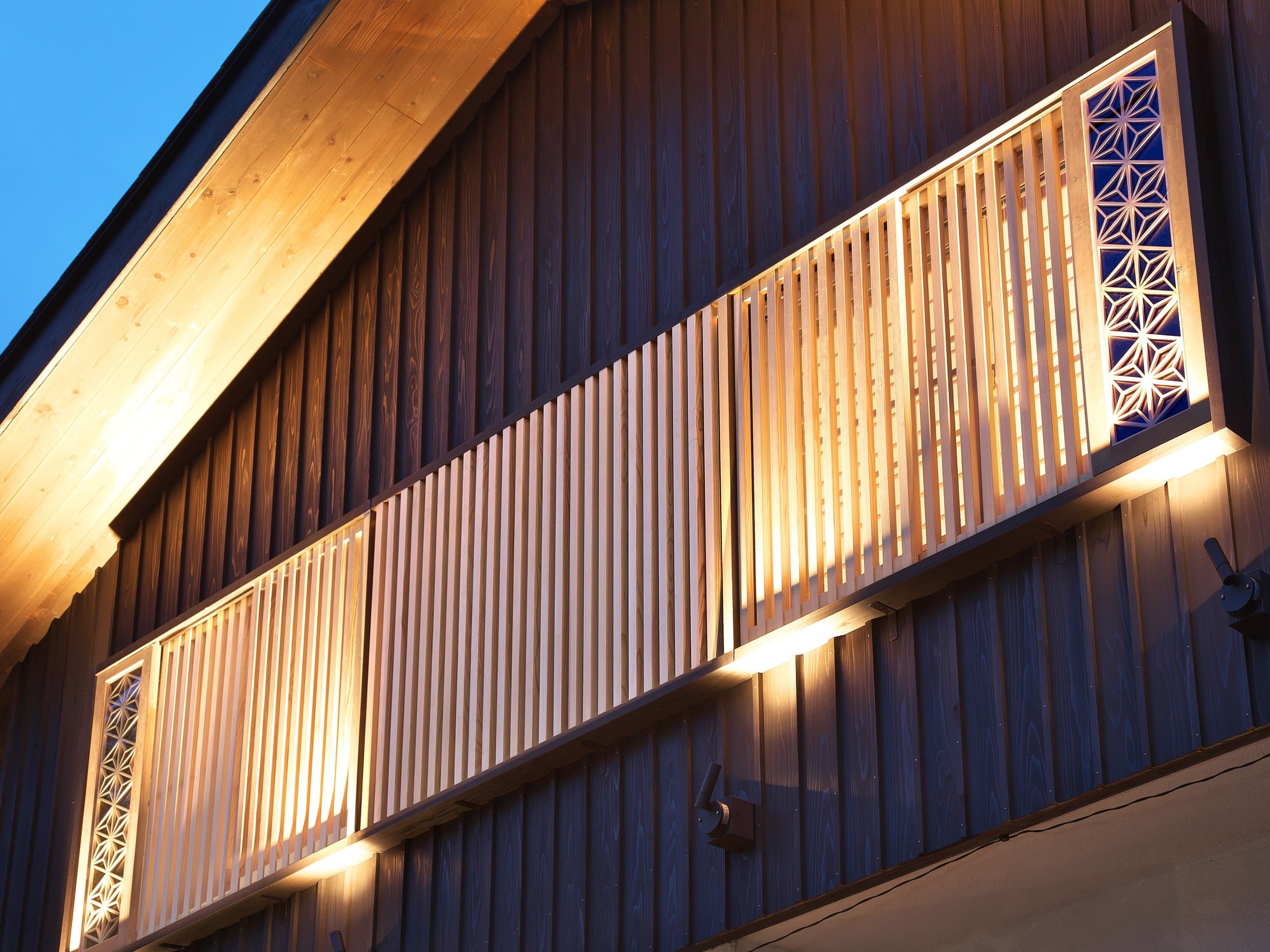 Architectural detail of wooden slat screens and asanoha lattice panels lit warmly against a dusk sky