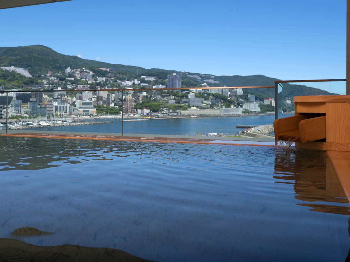 Rooftop open-air onsen bath with daytime sea view over Atami town and harbor