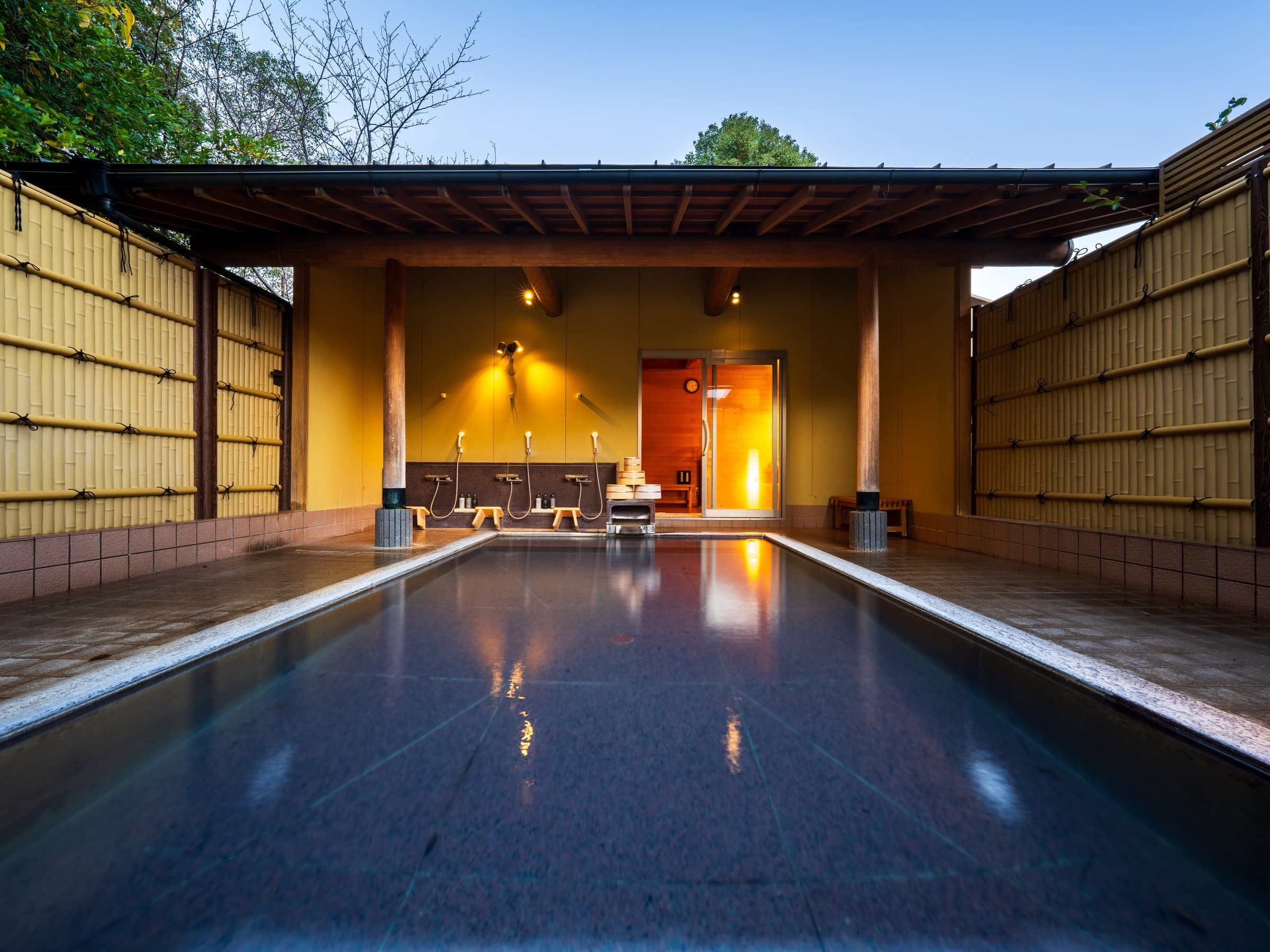 Spacious open-air rotenburo bath with bamboo fencing and timber roof glowing in evening twilight, Kappo Ryokan Kannawa Bettei, Beppu Onsen