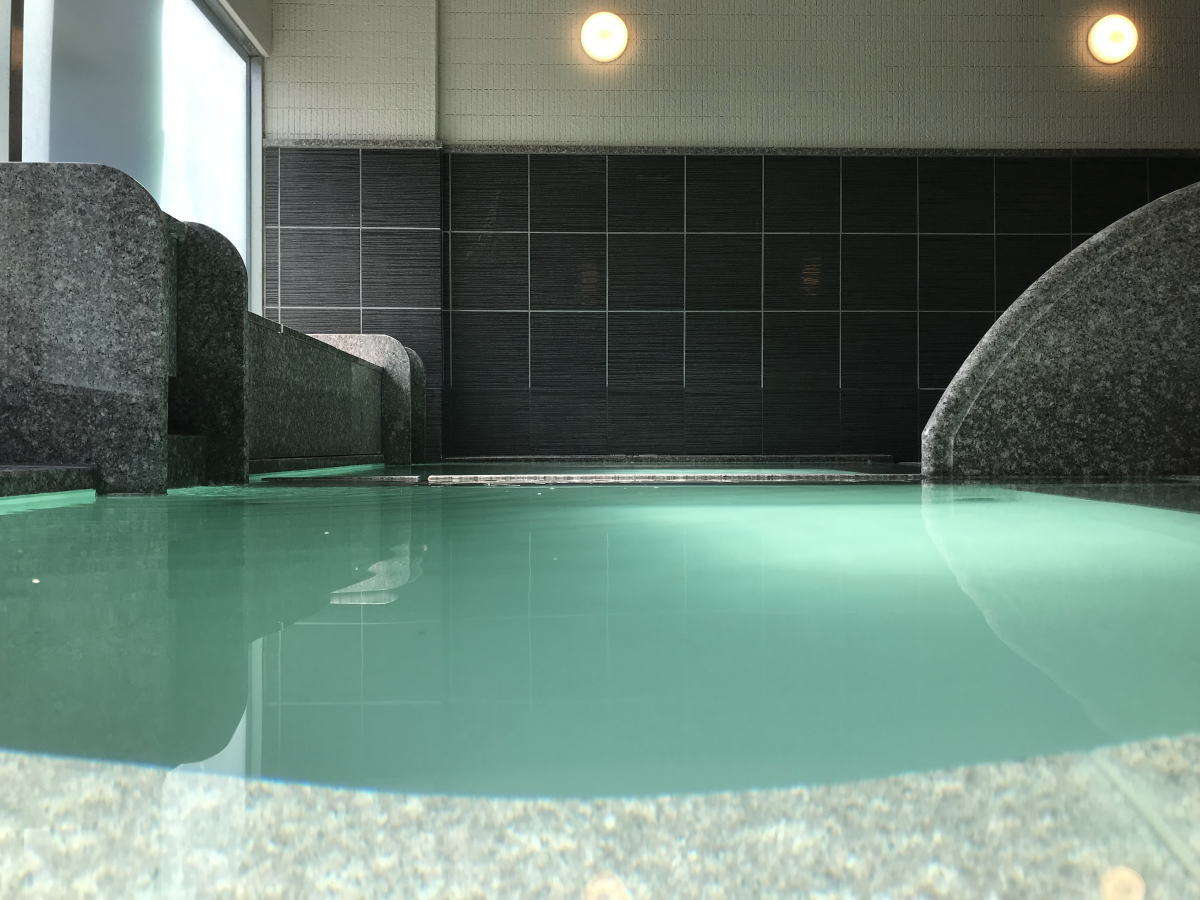 Large indoor onsen bath filled with milky green sulfur hot-spring water and dark tiled walls