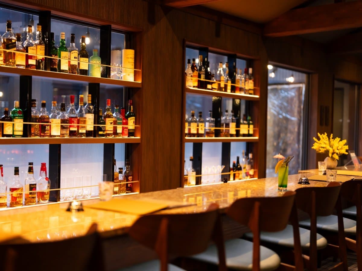 Warmly lit whisky bar counter backed by glass shelves of bottles in a wood-toned lounge