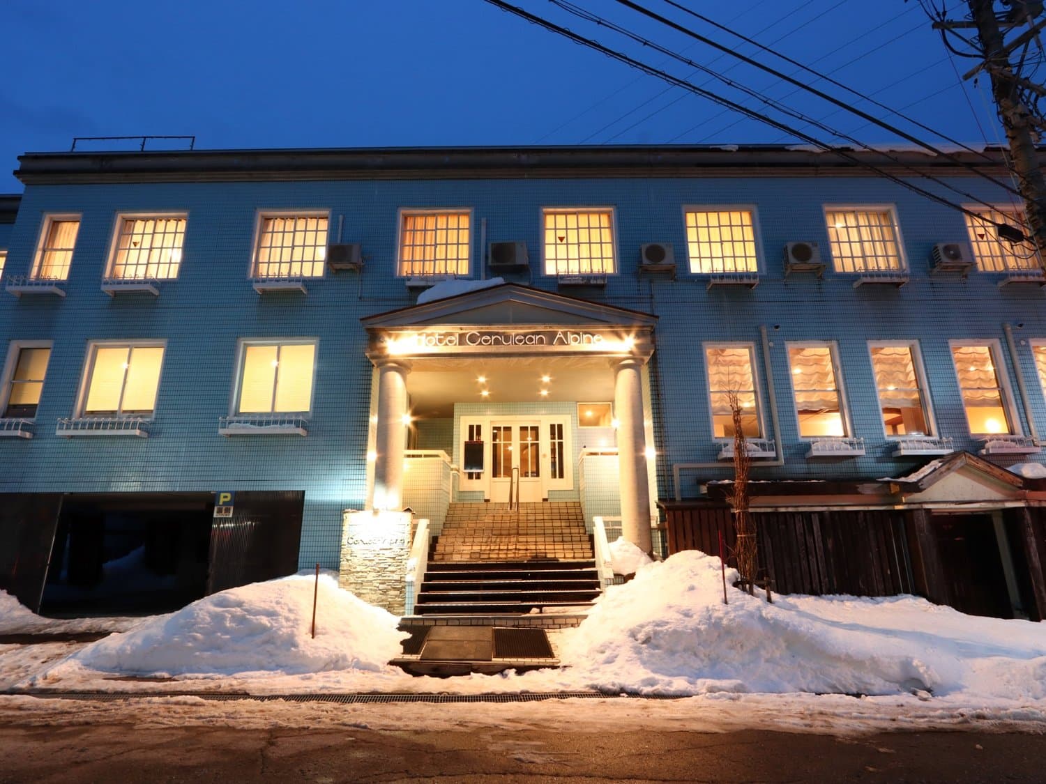Hotel Cerulean Alpen's blue-tiled main building lit at dusk amid winter snow, Tsugaike Onsen, Hakuba