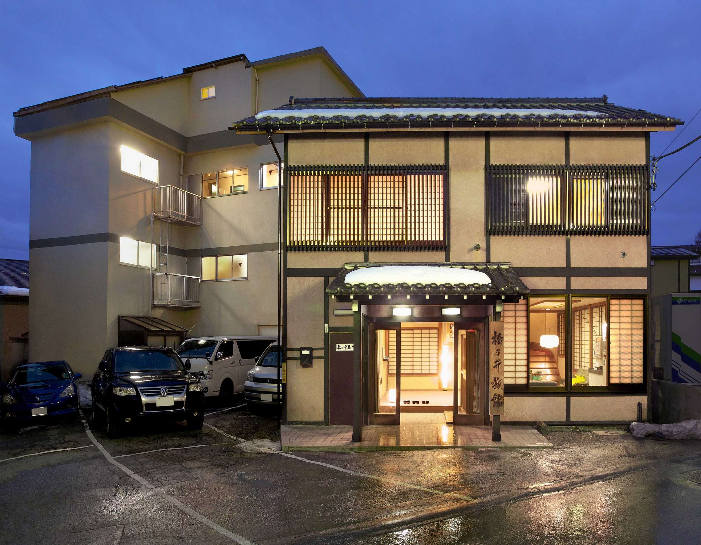 Three-story ryokan exterior at dusk with lit traditional latticed entrance and parking forecourt
