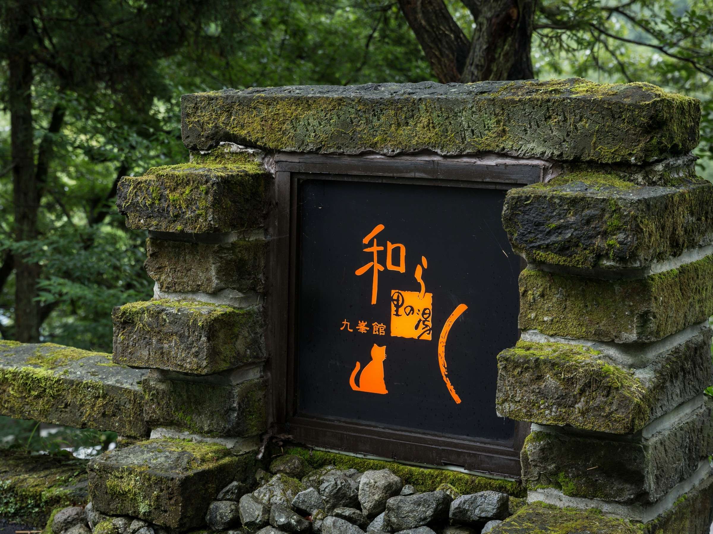 Mossy stone wall at Satonoyu Waraku with the inn's engraved logo plaque glowing amber against the forest