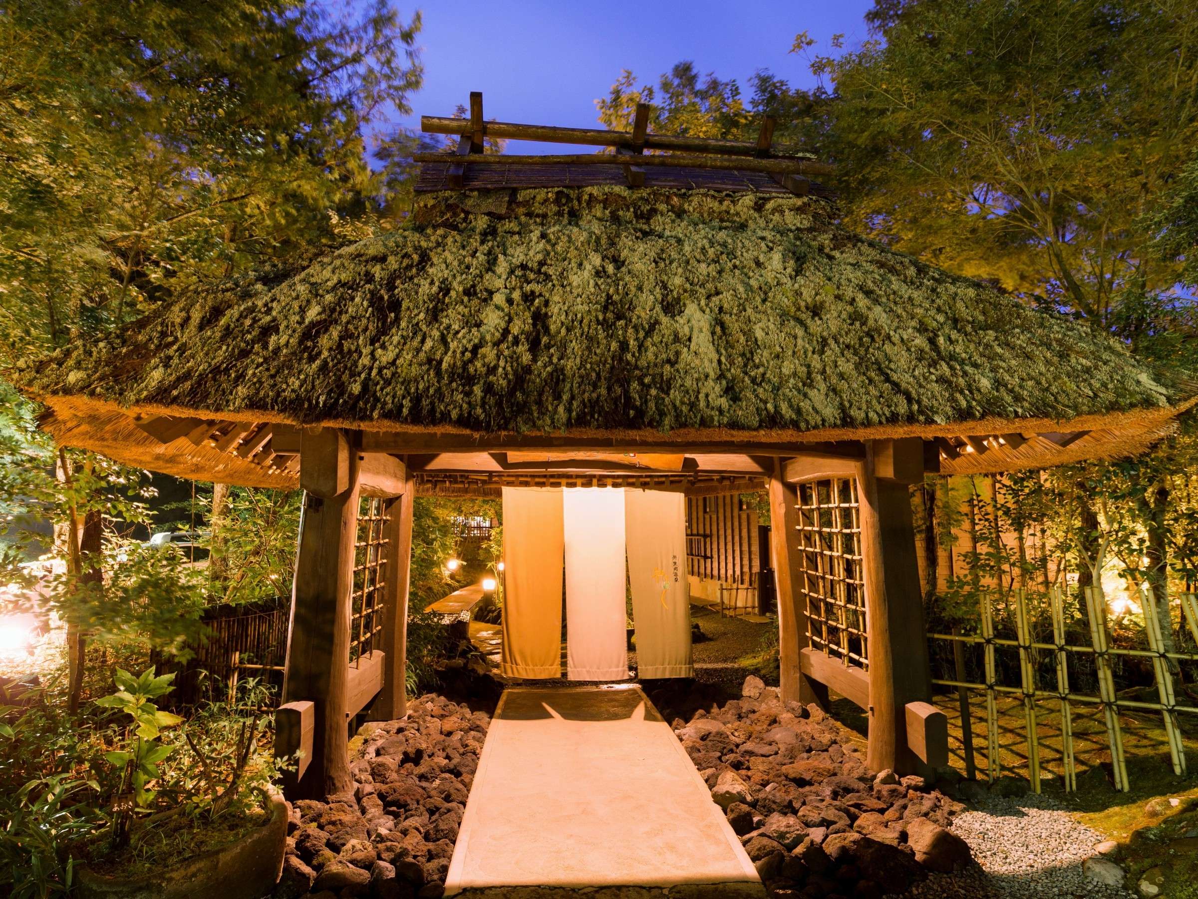 Satonoyu Waraku thatched-roof entrance gate illuminated at dusk, framed by greenery in Kurokawa Onsen