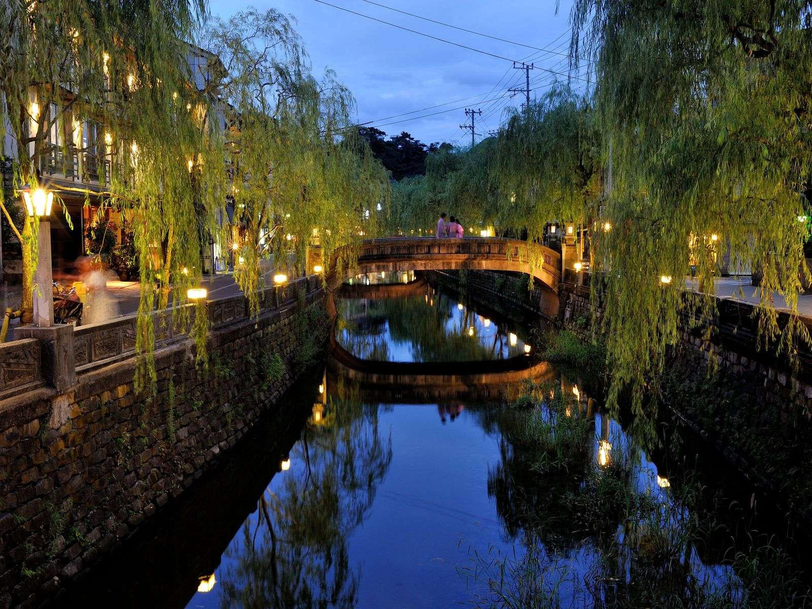 Willow-lined Kinosaki canal glowing with lanterns and a stone bridge at twilight, Yamamotoya, Kinosaki Onsen