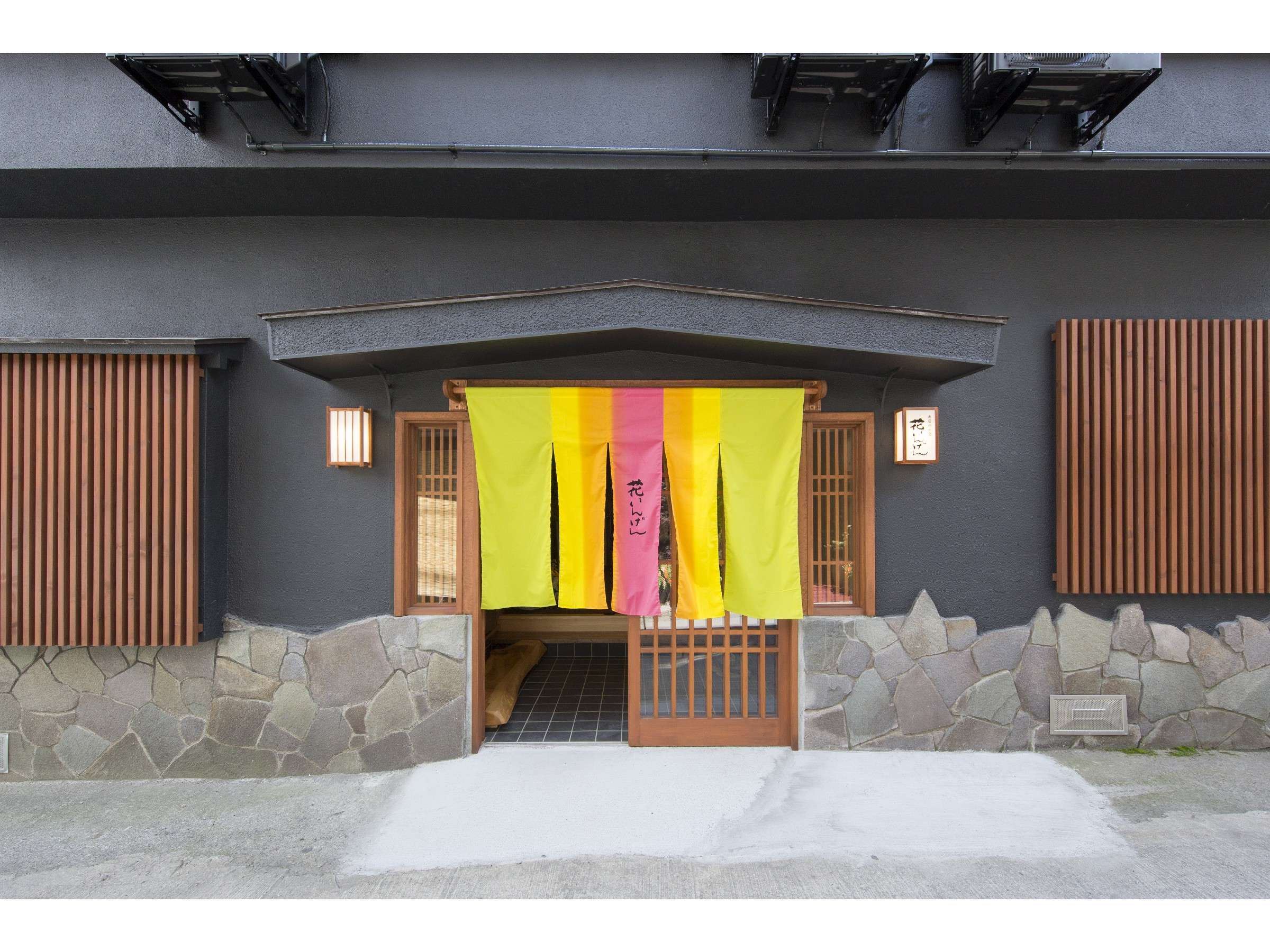Traditional inn entrance with dark facade, stone base and colorful gradient noren curtain over a wooden gate