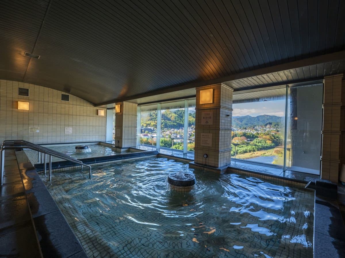 Spacious indoor hot-spring bath with mosaic-tiled floor and floor-to-ceiling windows framing the Ome mountains