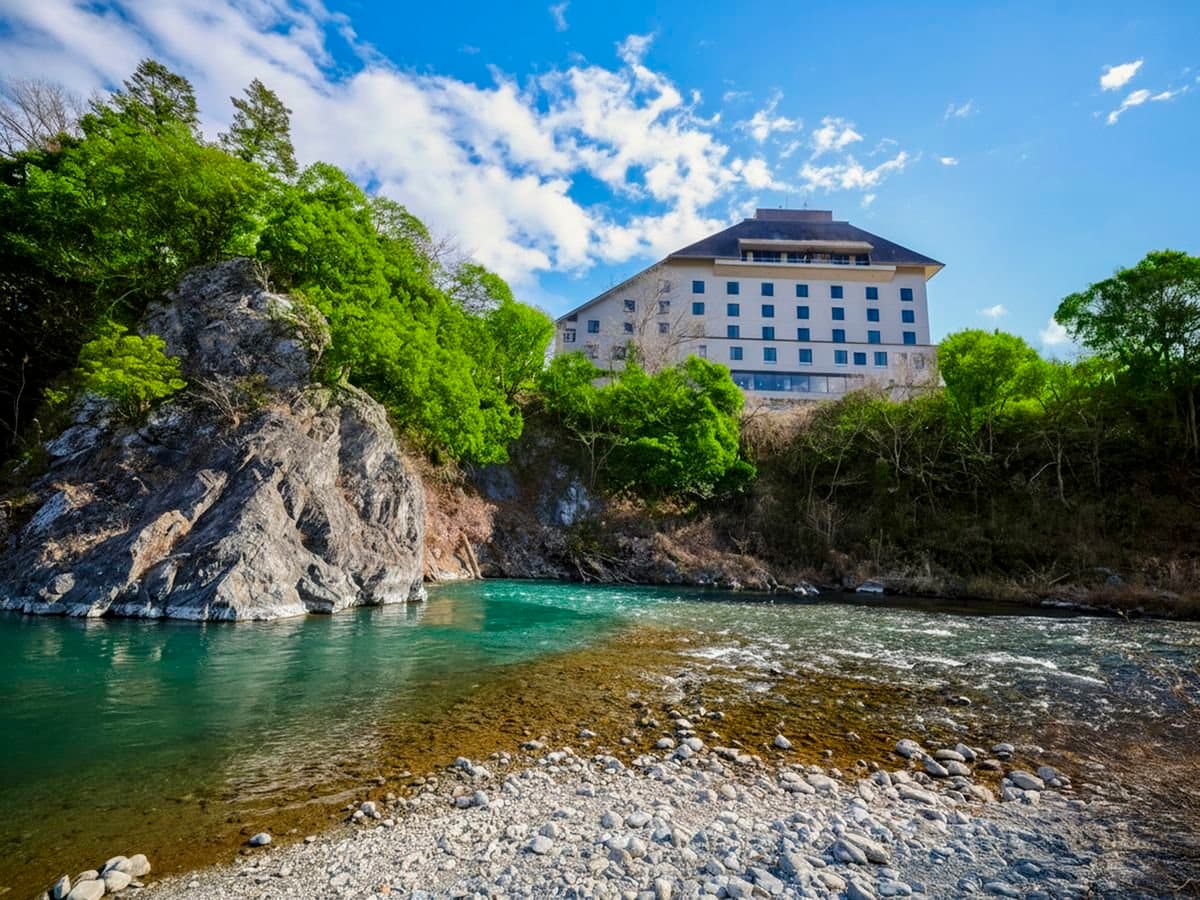 Kamenoi Hotel Ome rising above the emerald Tama River gorge under a blue sky in western Tokyo's mountains