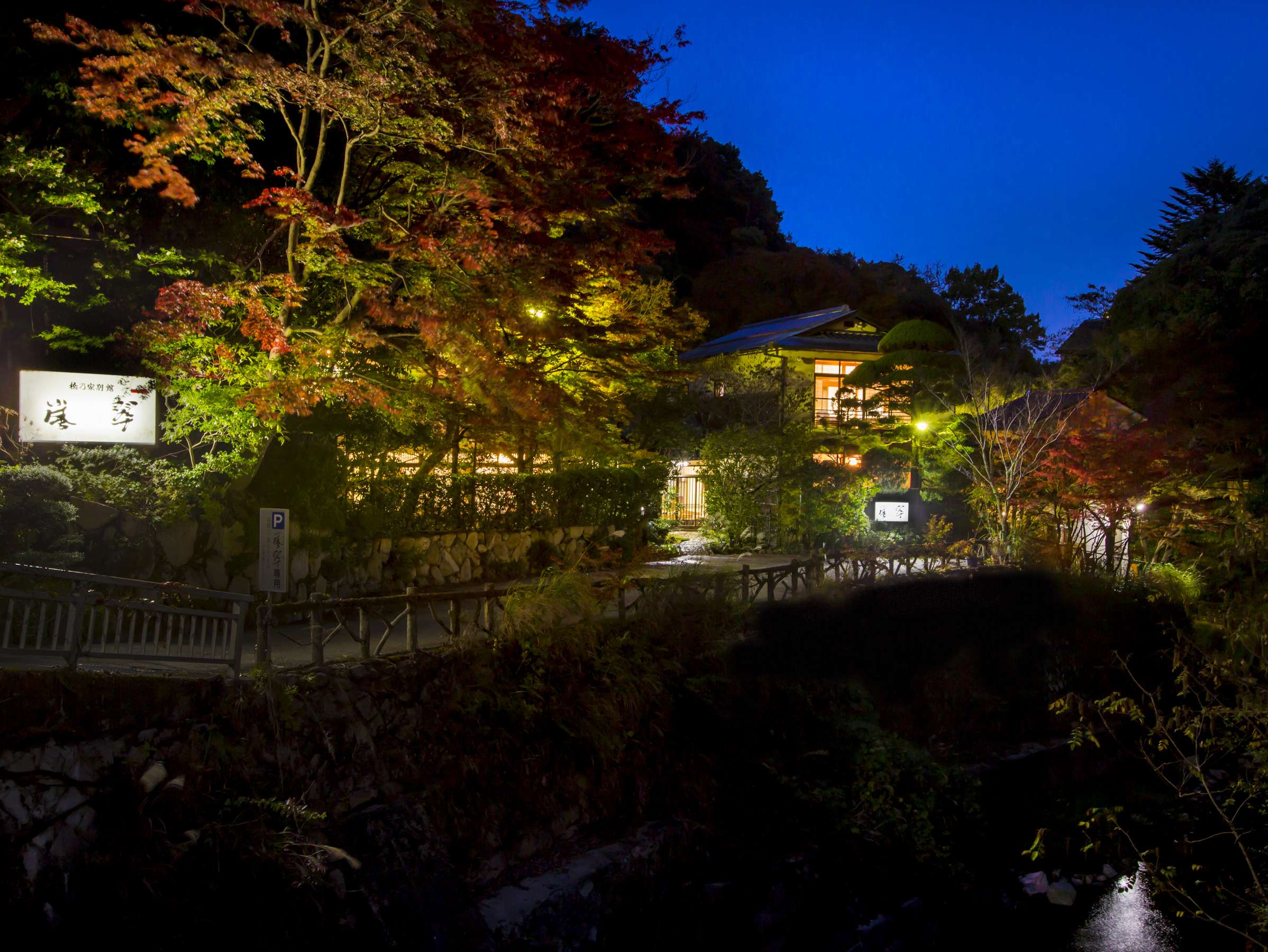 Hashinoya Bekkan Ransui's Meiji-era villa lit at blue-hour dusk among autumn maples, with its calligraphy signboard and a stone bridge over the Arima stream in the foreground