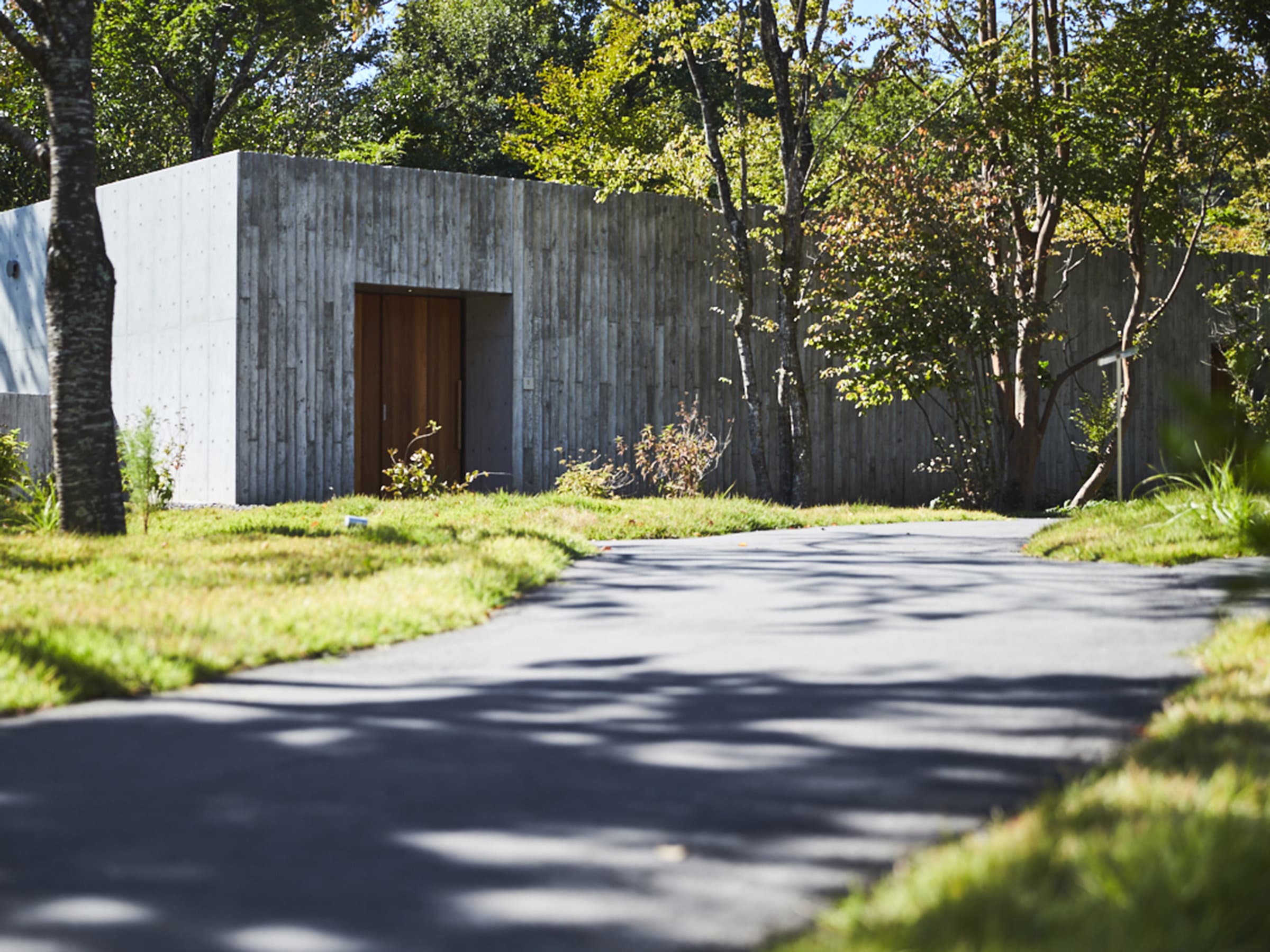 Sanso Warabino concrete-and-timber villa entrance with a curving garden path among trees in Yufuin, Oita