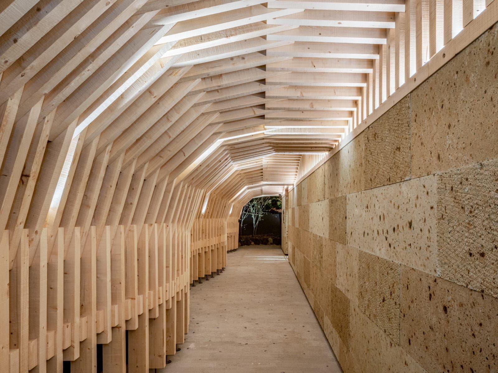 Sculptural corridor lined with fanned wooden ribs and a textured stone wall leading to a courtyard