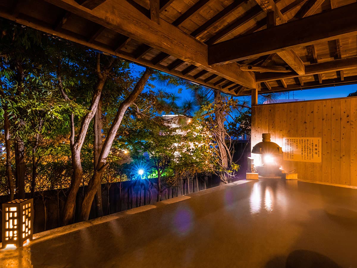 Open-air rotenburo bath at night framed by trees, timber roof and a glowing stone lantern, Ryotei Matsubaya, Beppu Onsen