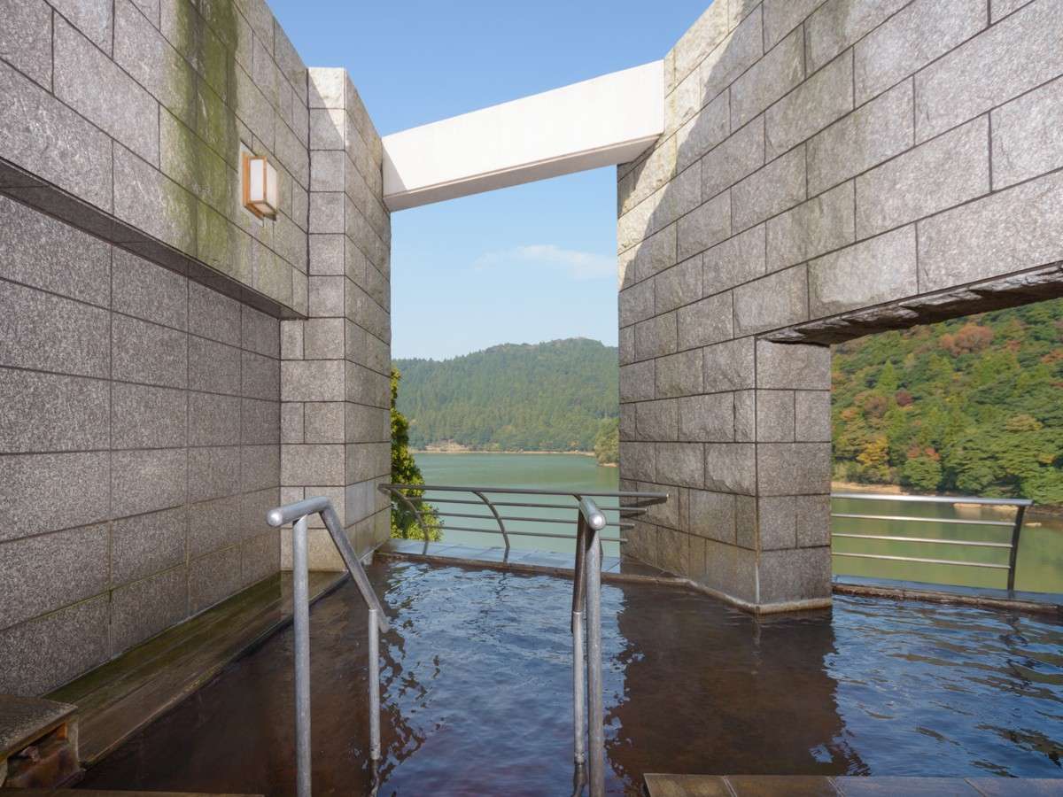 Open-air stone rotenburo bath at Unzen Azumaen framing a view across Lake Shirakumo to forested hills