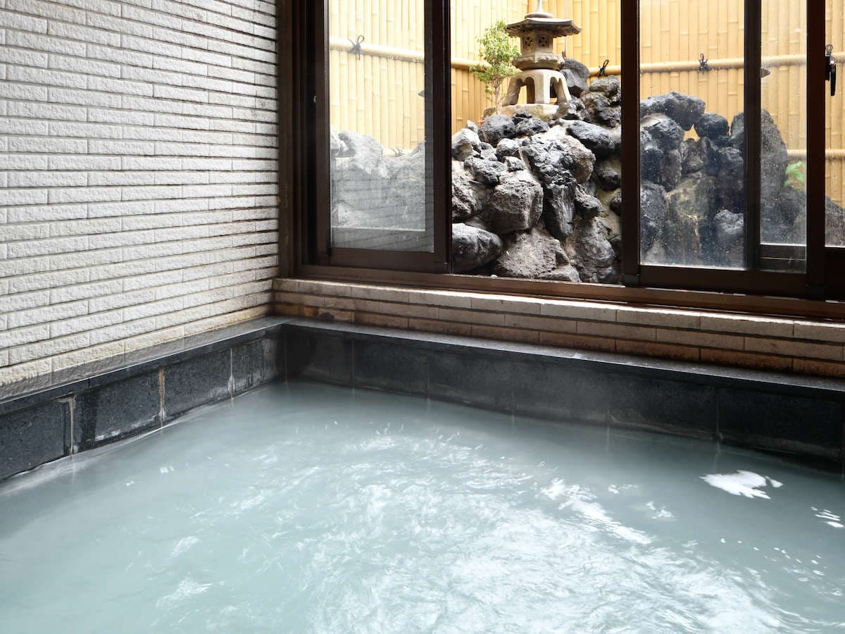 Indoor hot-spring bath at Ryokan Tamura filled with milky Jizo-no-Yu spring water, with a stone lantern garden visible through the window, Kusatsu Onsen