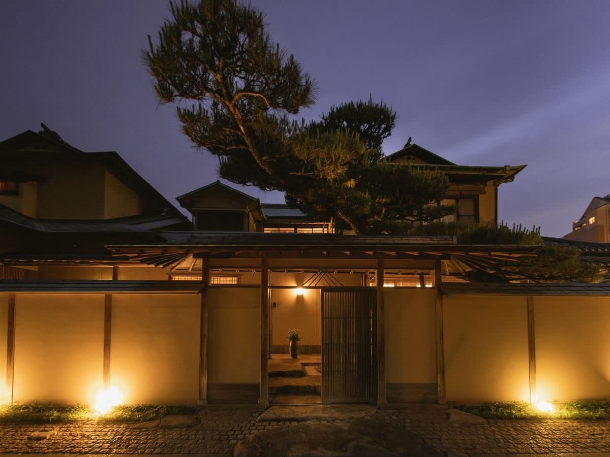 Dogo Onsen Ryotei Umenoya sukiya-style inn entrance gate and tiled roofs lit warmly at dusk under a pine tree