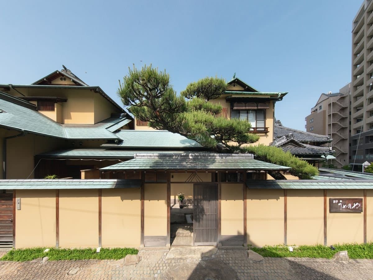 Daytime exterior of Umenoya in Dogo Onsen with green-copper tiled roofs and a sculpted pine over the gated earthen wall