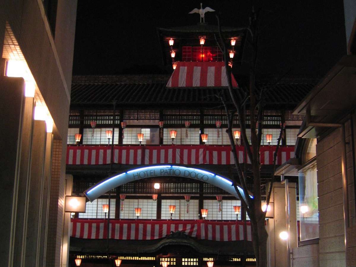 Dogo Onsen Honkan bathhouse glowing at night framed by the Hotel Patio Dogo entrance arch