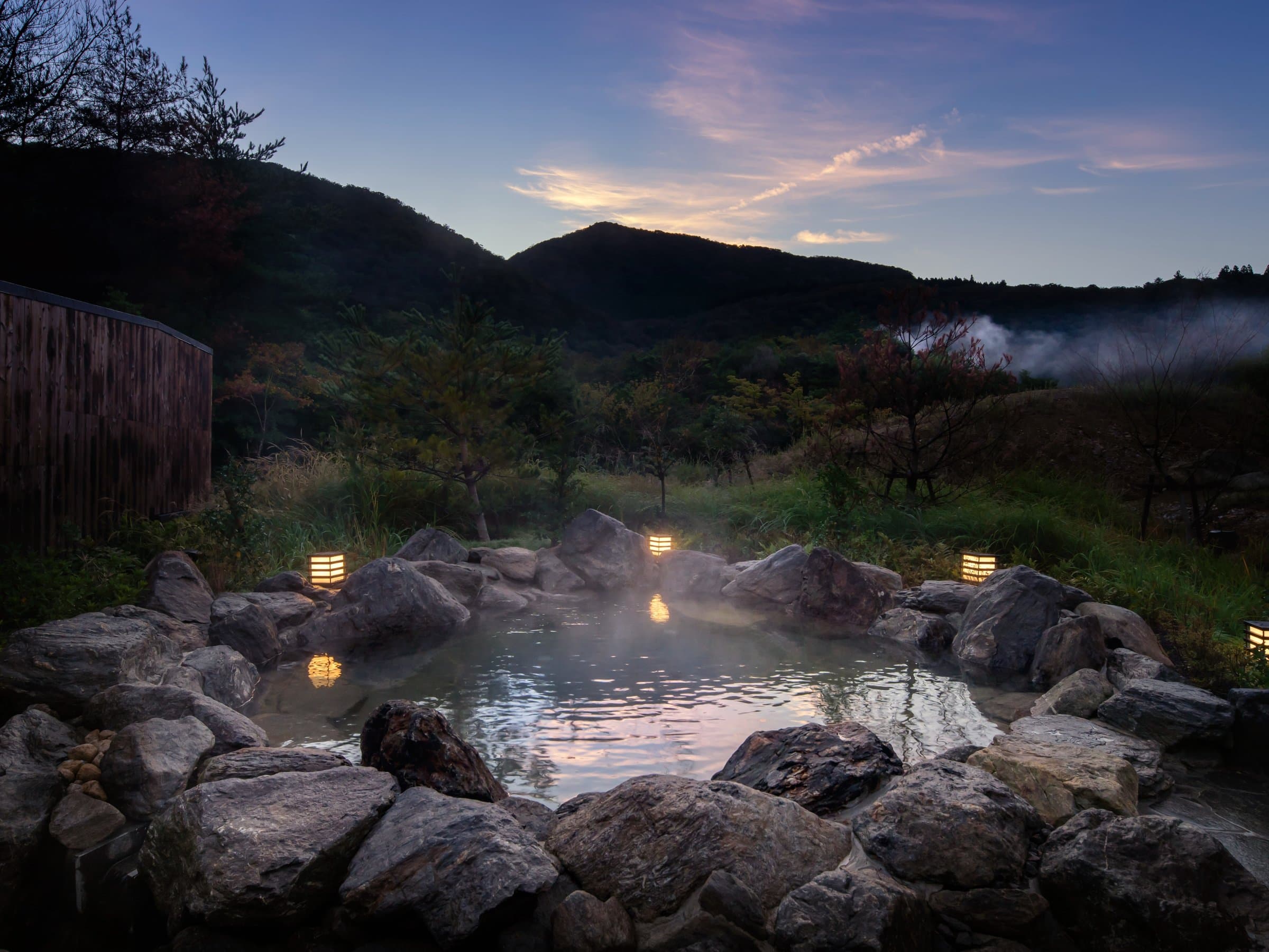 Rock-rimmed open-air rotenburo bath at KAI Unzen glowing at dusk with steam rising over the Unzen valley and distant hot-spring vapor