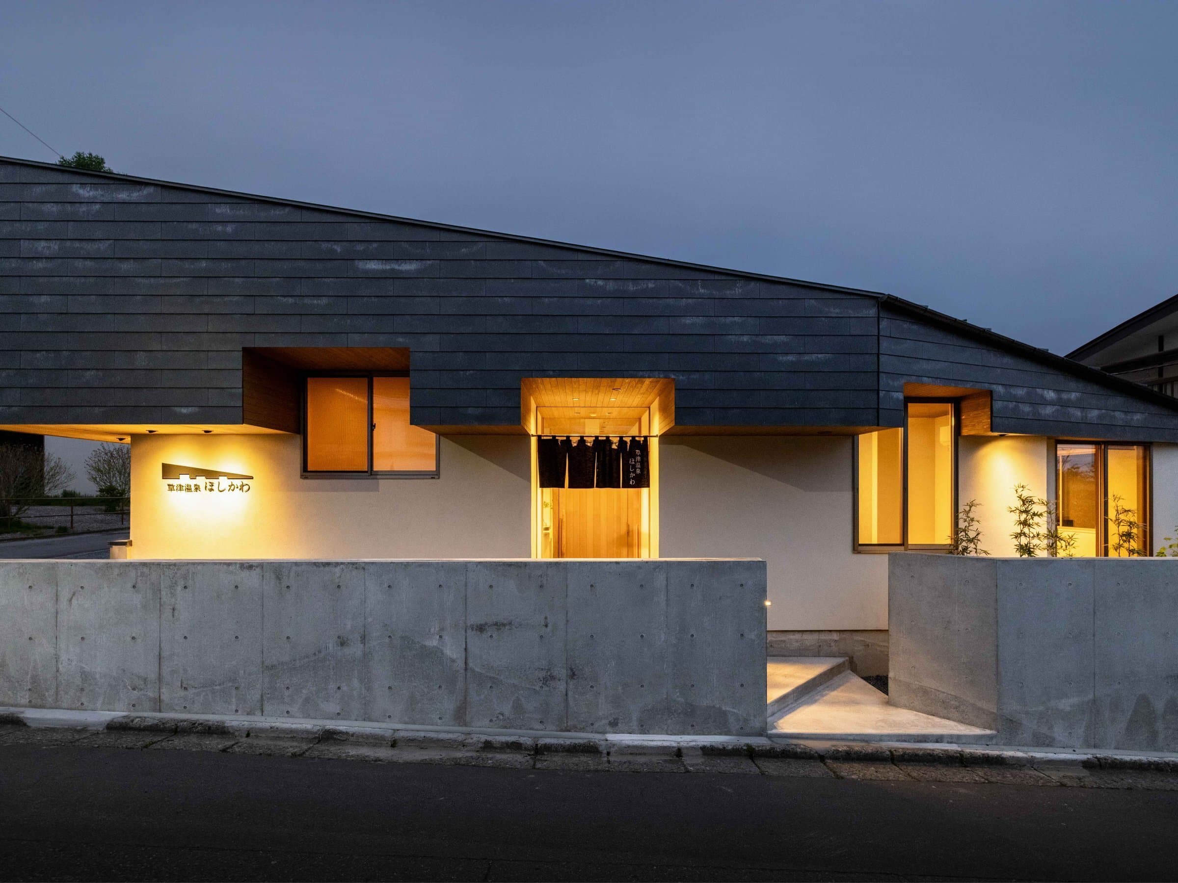 Kusatsu Onsen Hoshikawa's modern single-storey ryokan exterior lit at dusk with warm entrance lighting and on-site signage
