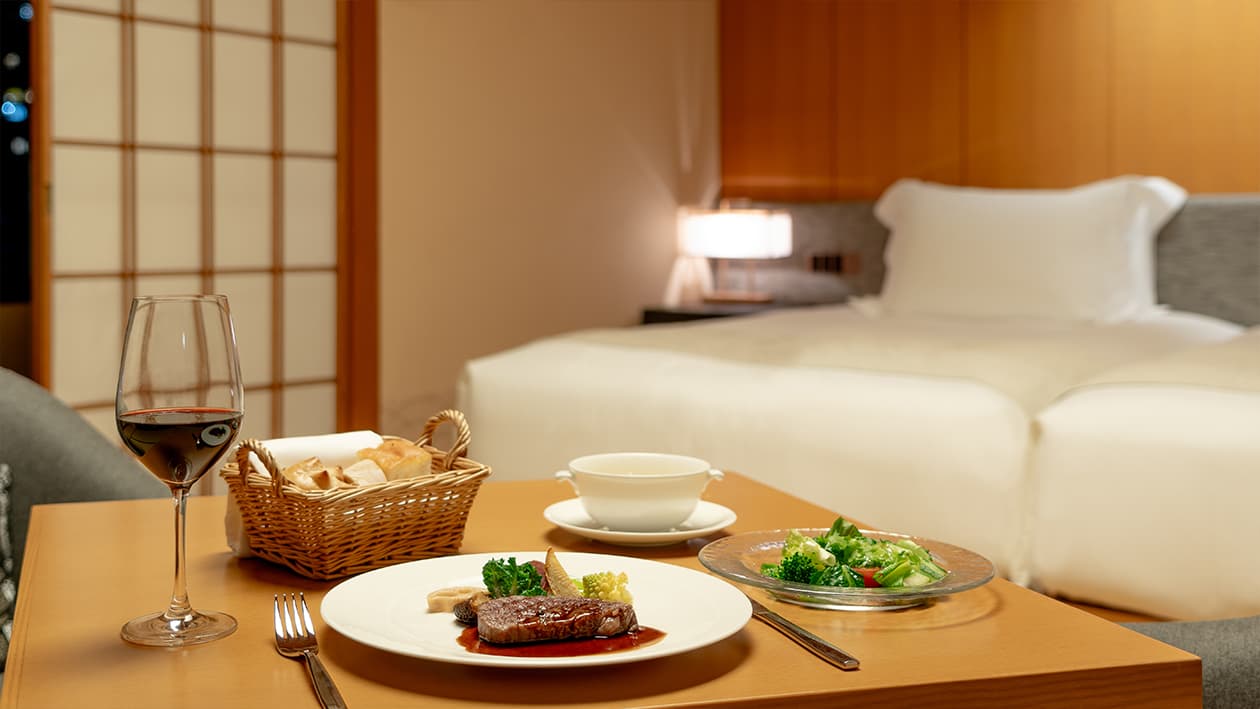 In-room dining with grilled beef, soup and red wine served beside a guest bed at Takanawa Hanakohro Tokyo