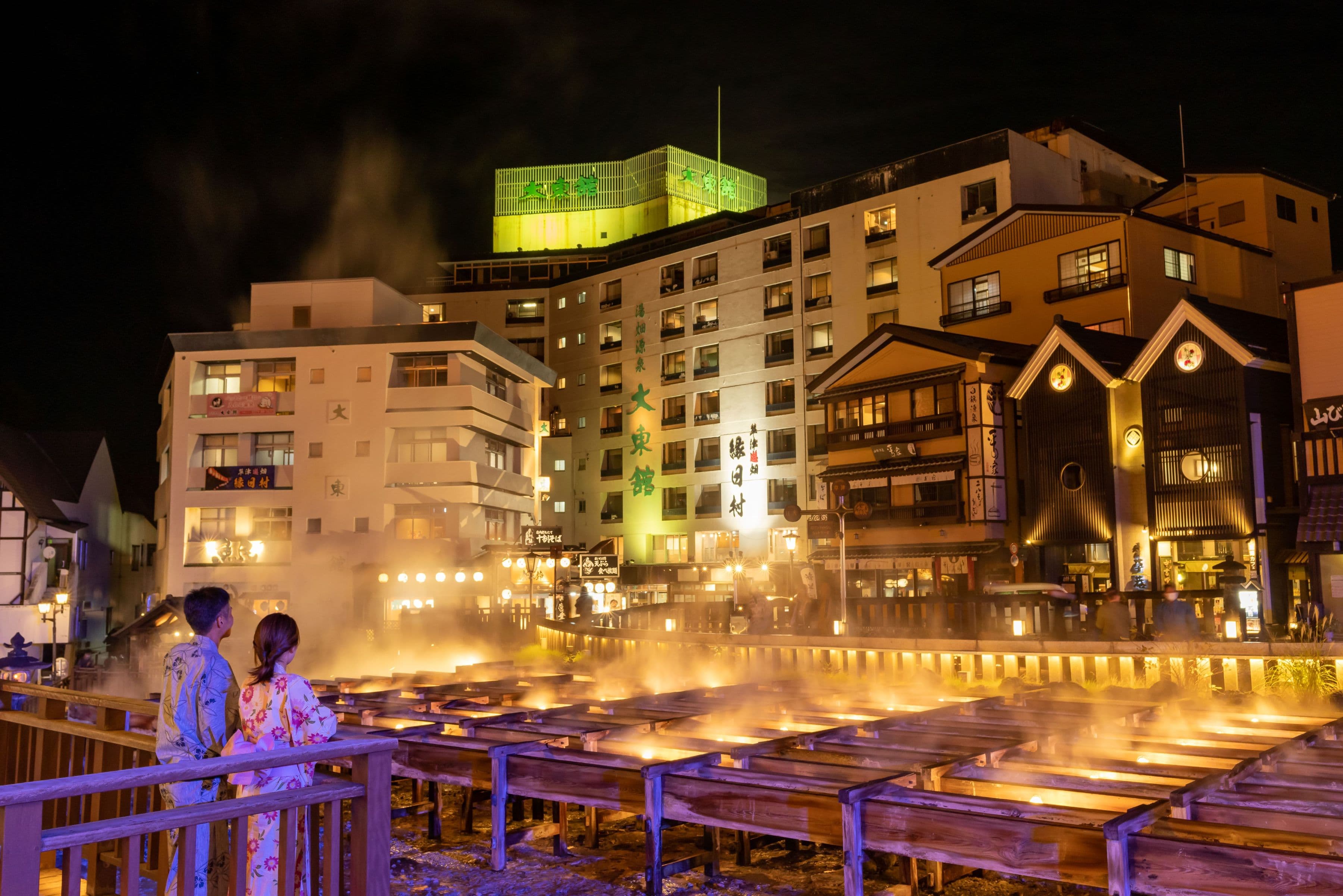 Illuminated hot-spring building overlooking the steaming Yubatake field at night in Kusatsu Onsen