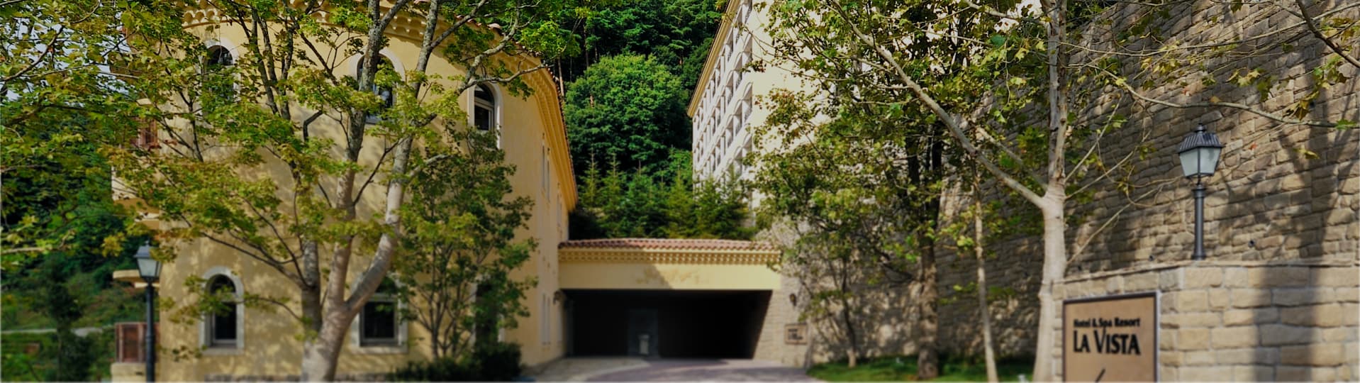 Exterior of La Vista Fuji Kawaguchiko, a Mediterranean-style stone-and-stucco resort hotel set among trees with its stone entrance plaque