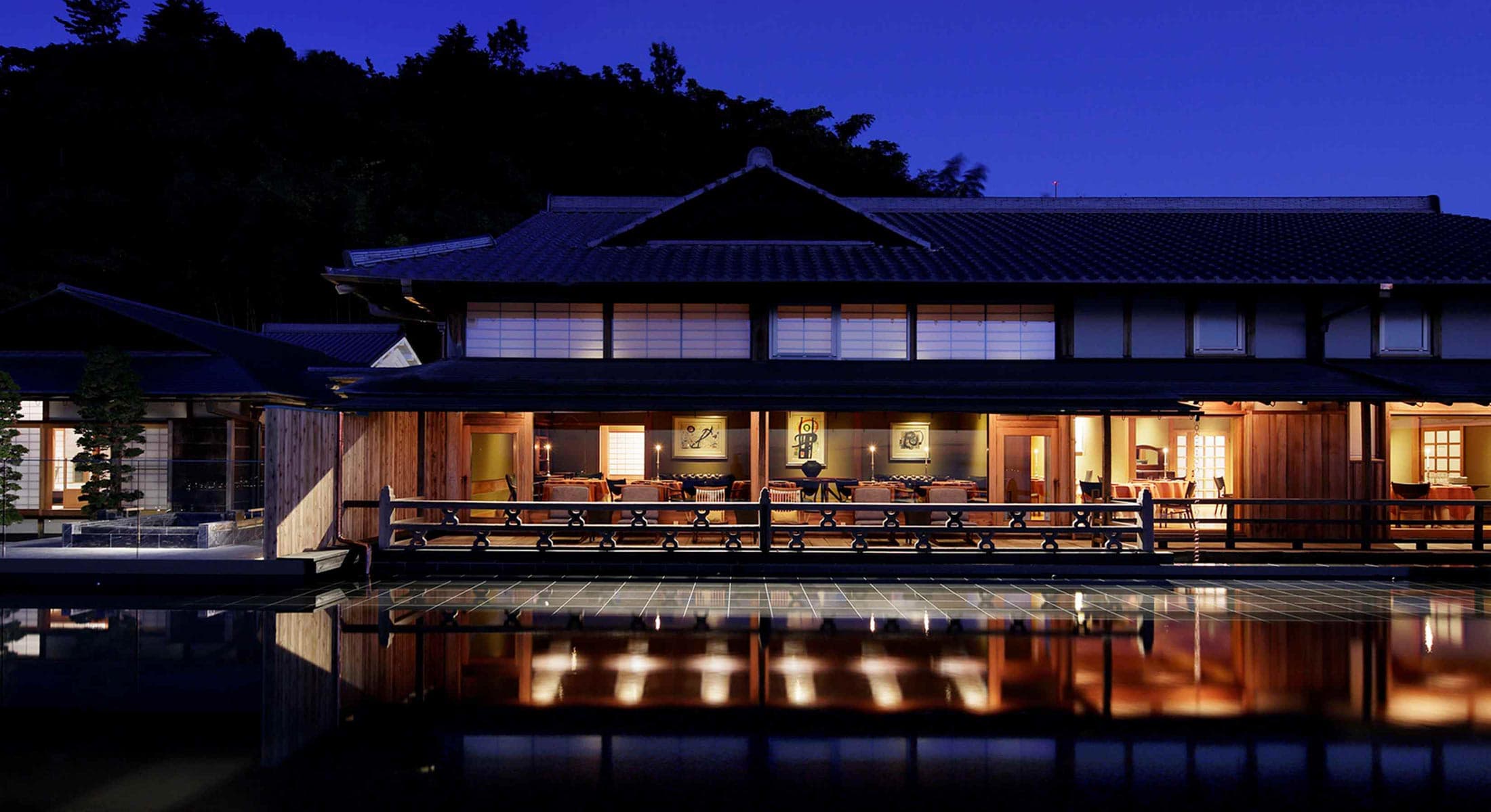 Illuminated open lounge of the Hiramatsu Atami at night, glowing shoji and a wide veranda over a still water terrace