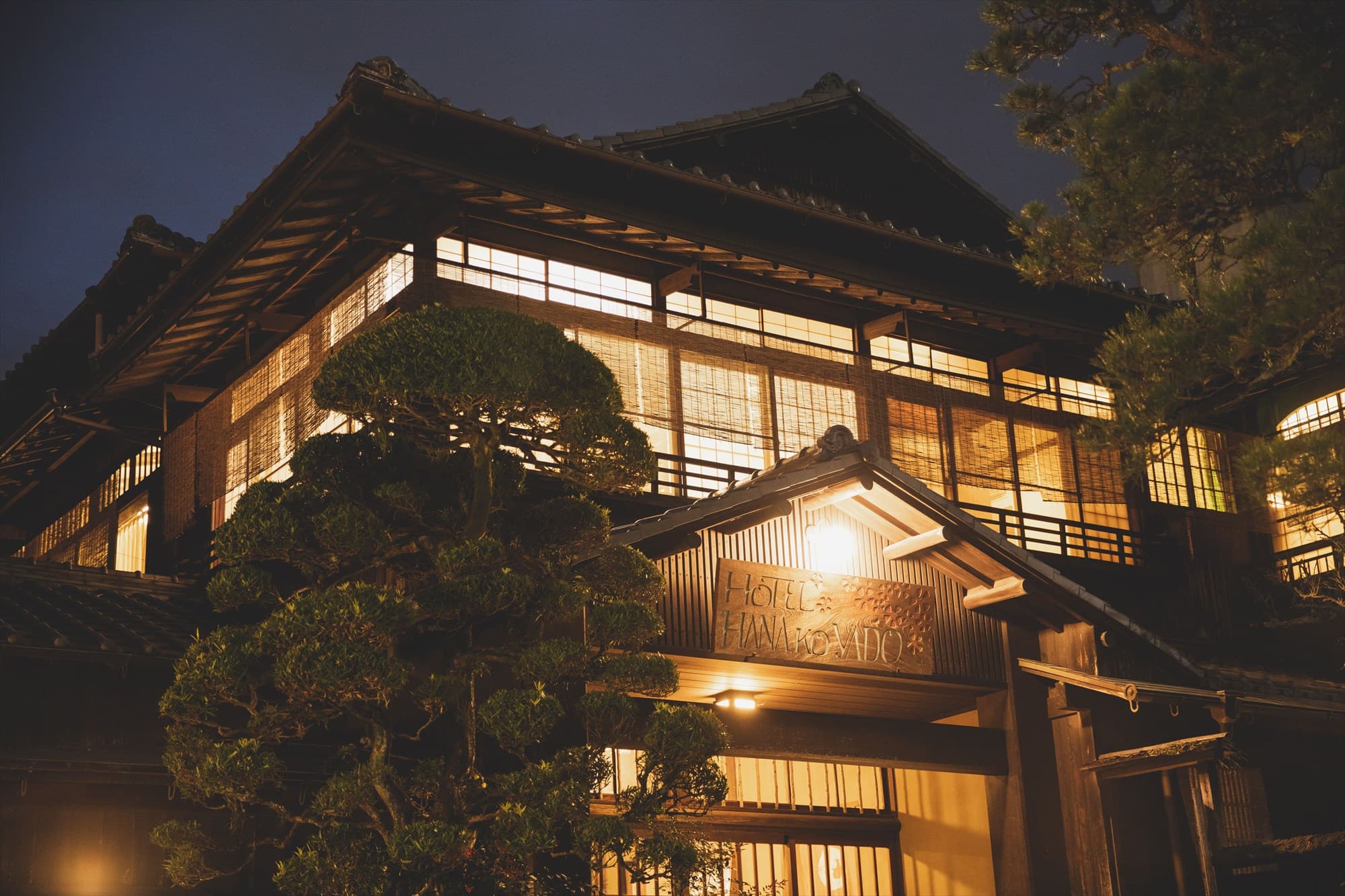 Hotel Hanakoyado historic wooden ryokan exterior illuminated at dusk with lit lantern entrance and pine trees, Arima Onsen
