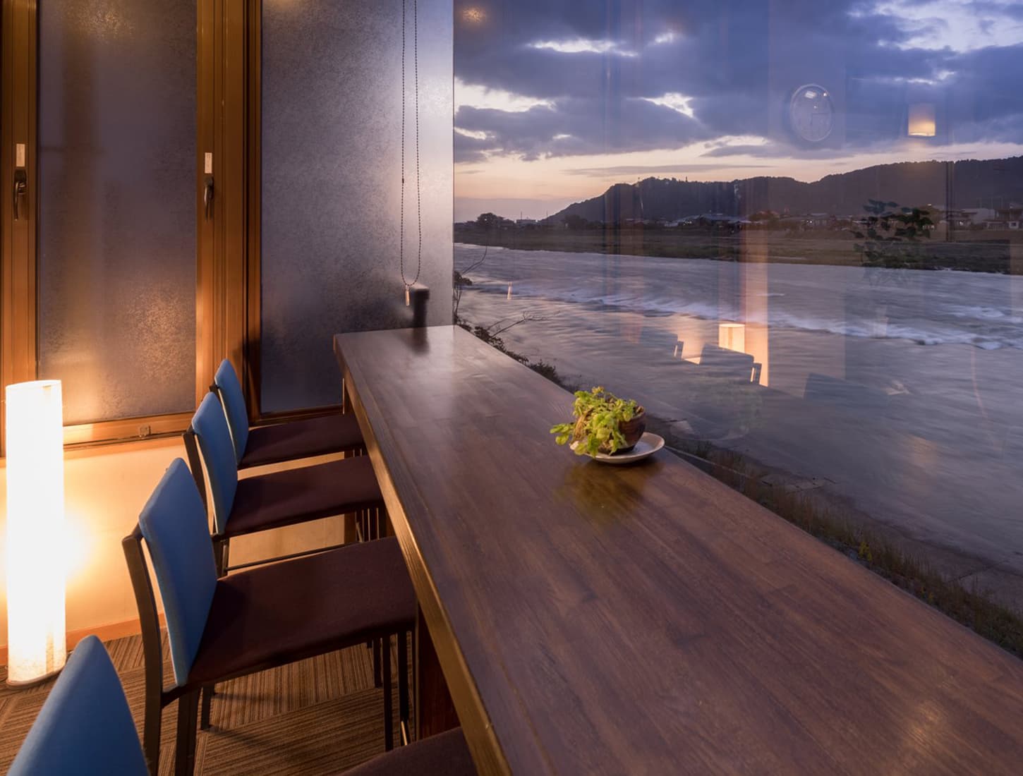 River-view counter lounge at Kiyonoya at dusk with a long wood table, floor lamp and the Chikugo River through floor-to-ceiling glass