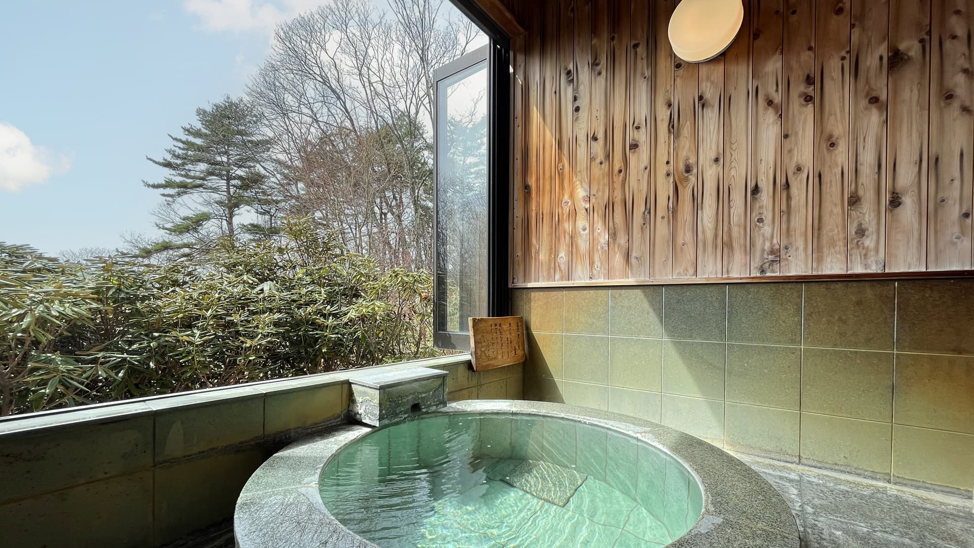 Round stone private kashikiri bath with milky-green hot spring water, cedar wall and a large window onto trees at Vent Vert