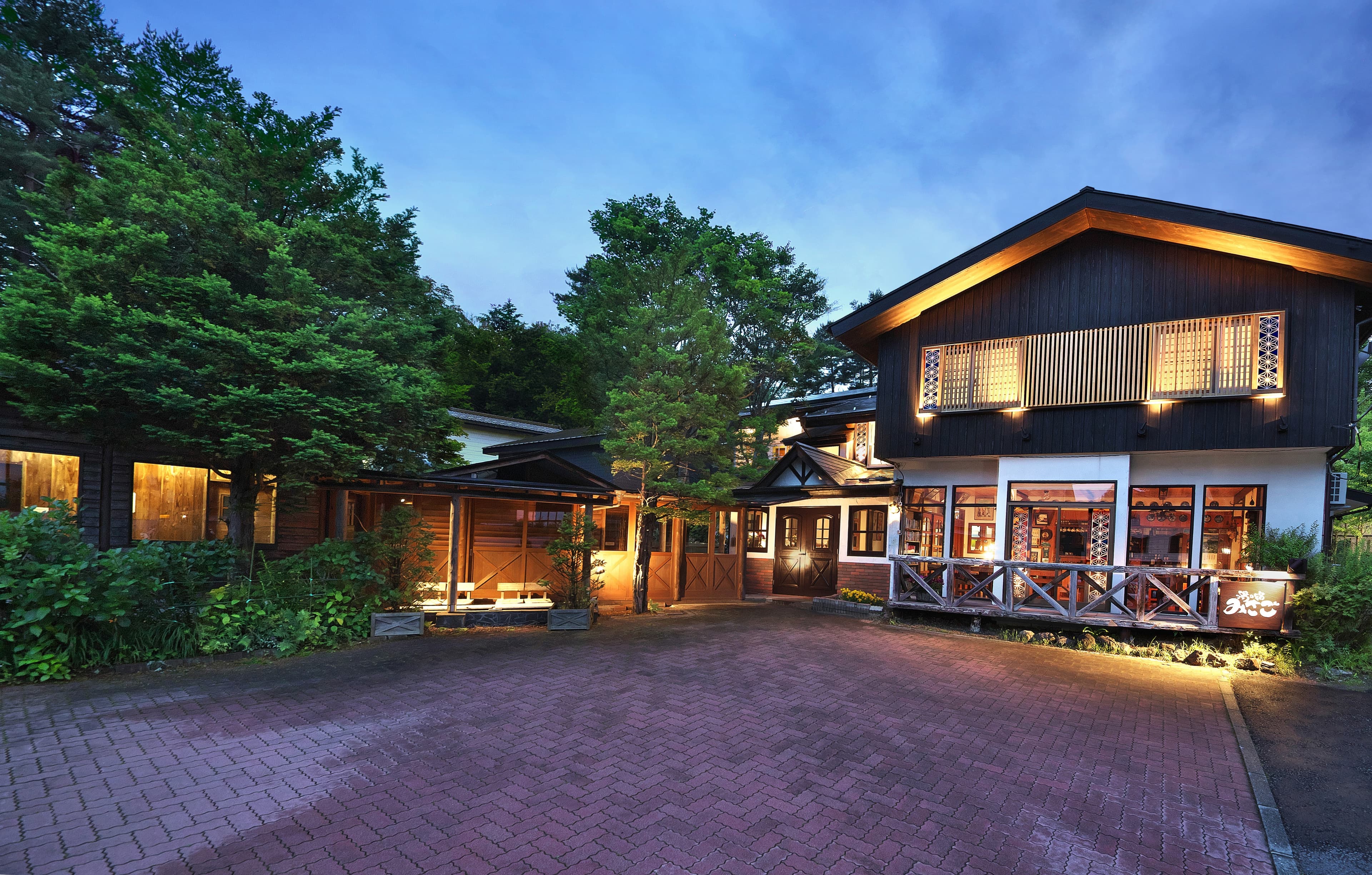 Wood-clad mountain inn lit warmly at dusk with a brick forecourt and surrounding pines in summer